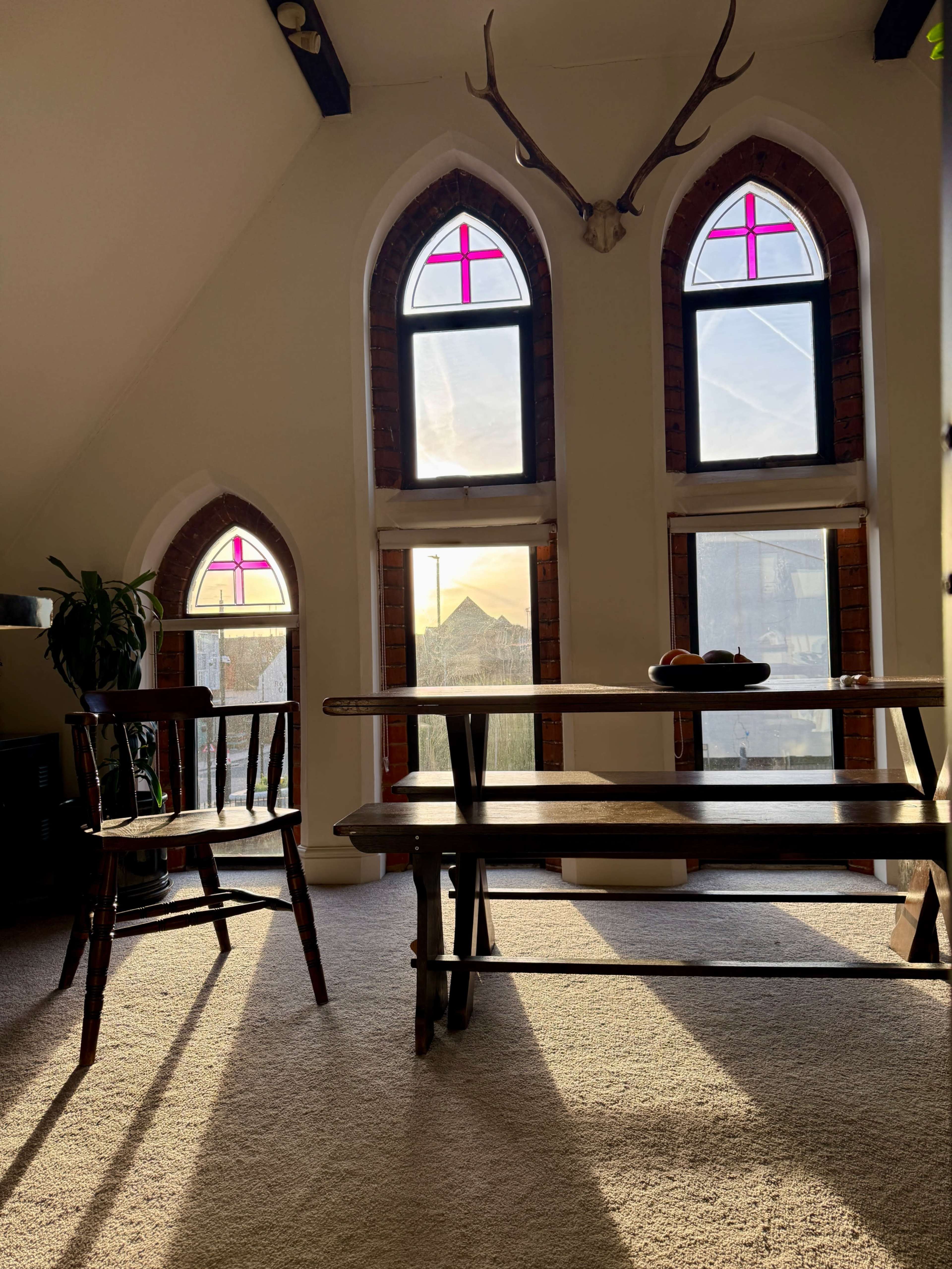 The scene depicts a cozy room with arched windows featuring pink stained glass, a wooden table, a chair, and soft sunlight casting shadows on the carpet.