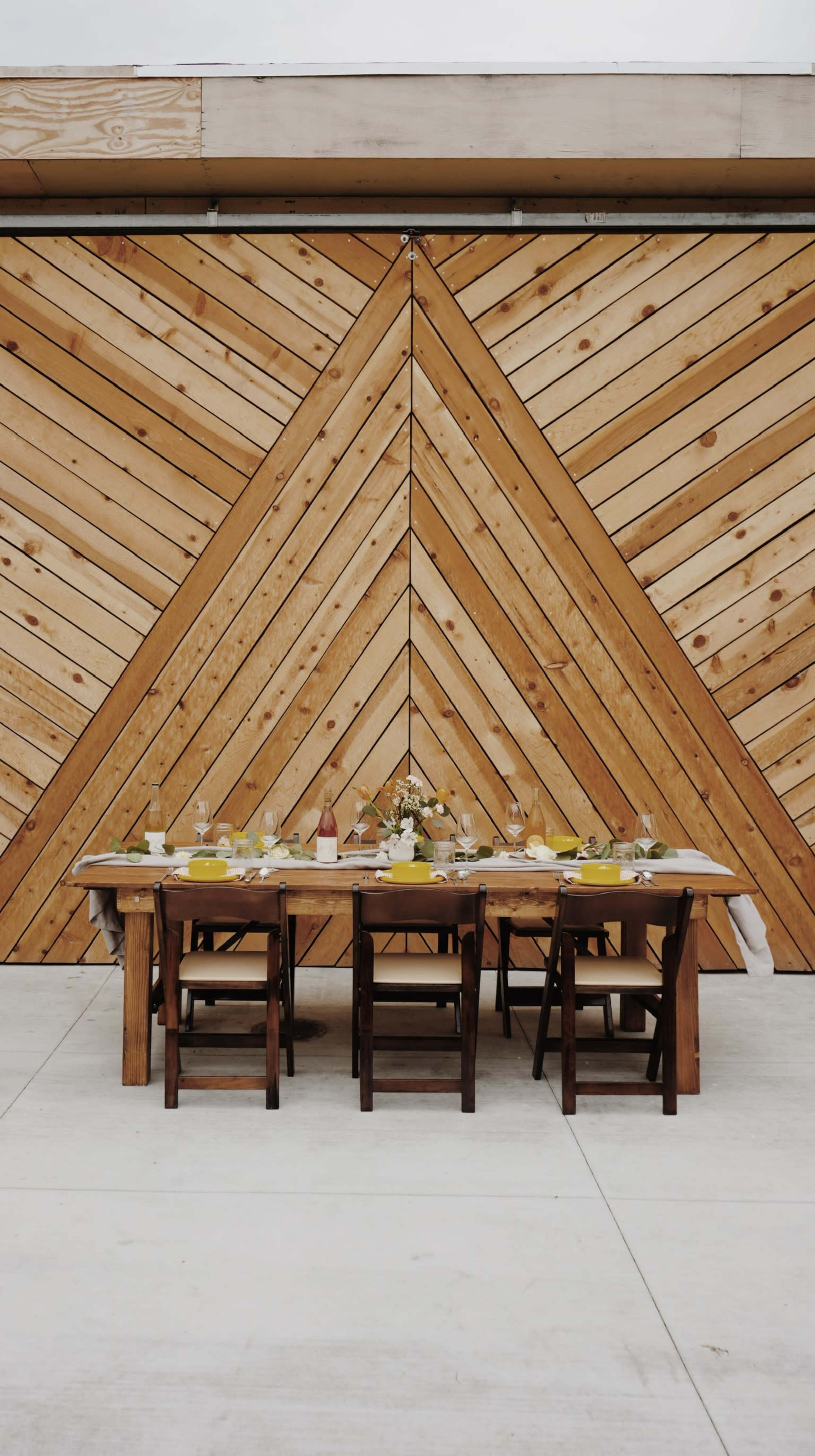A long wooden table is set for a meal in front of a large, chevron-patterned wooden wall.