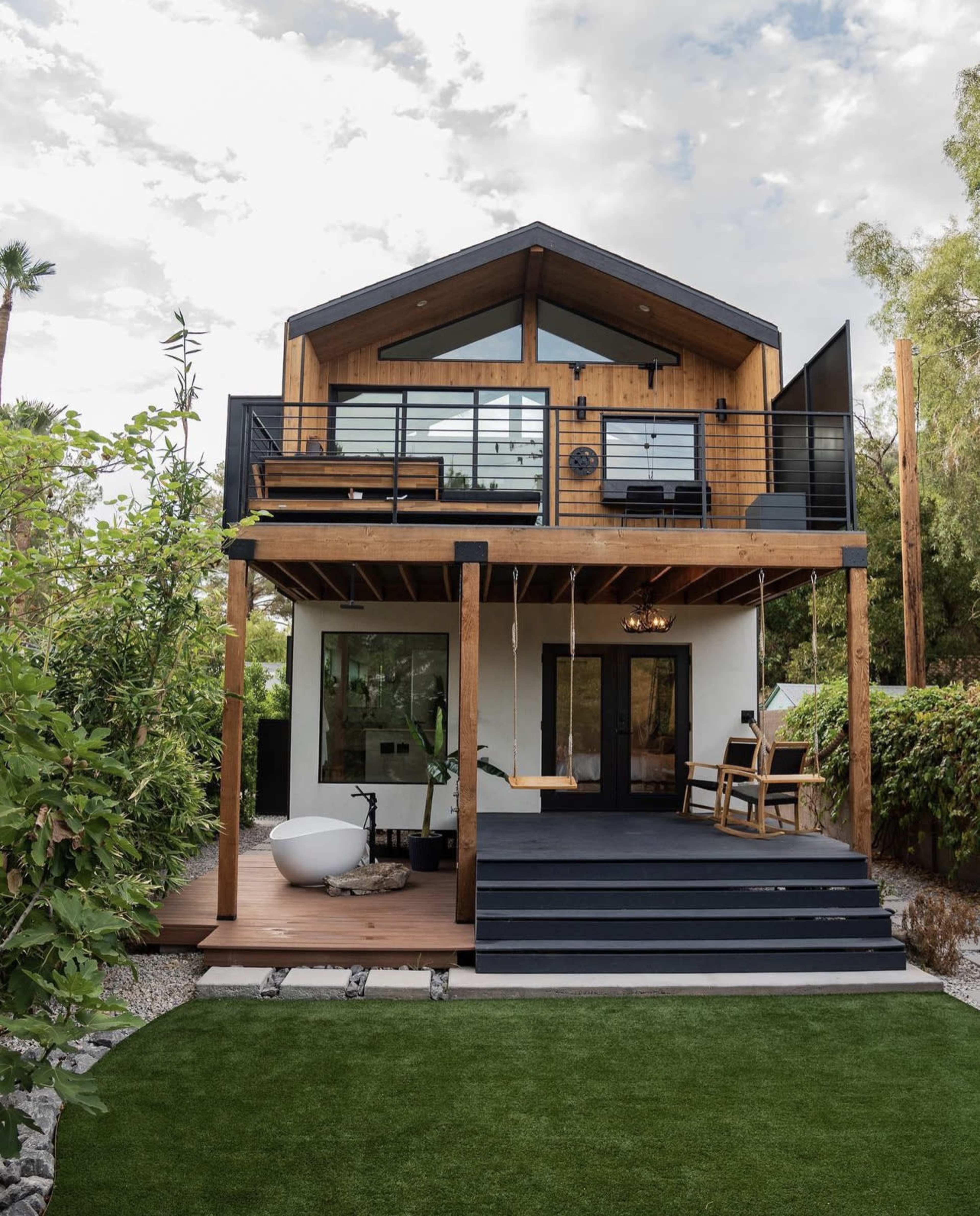 A modern two-story house features a wooden facade, large windows, and a balcony, surrounded by greenery and a landscaped yard.