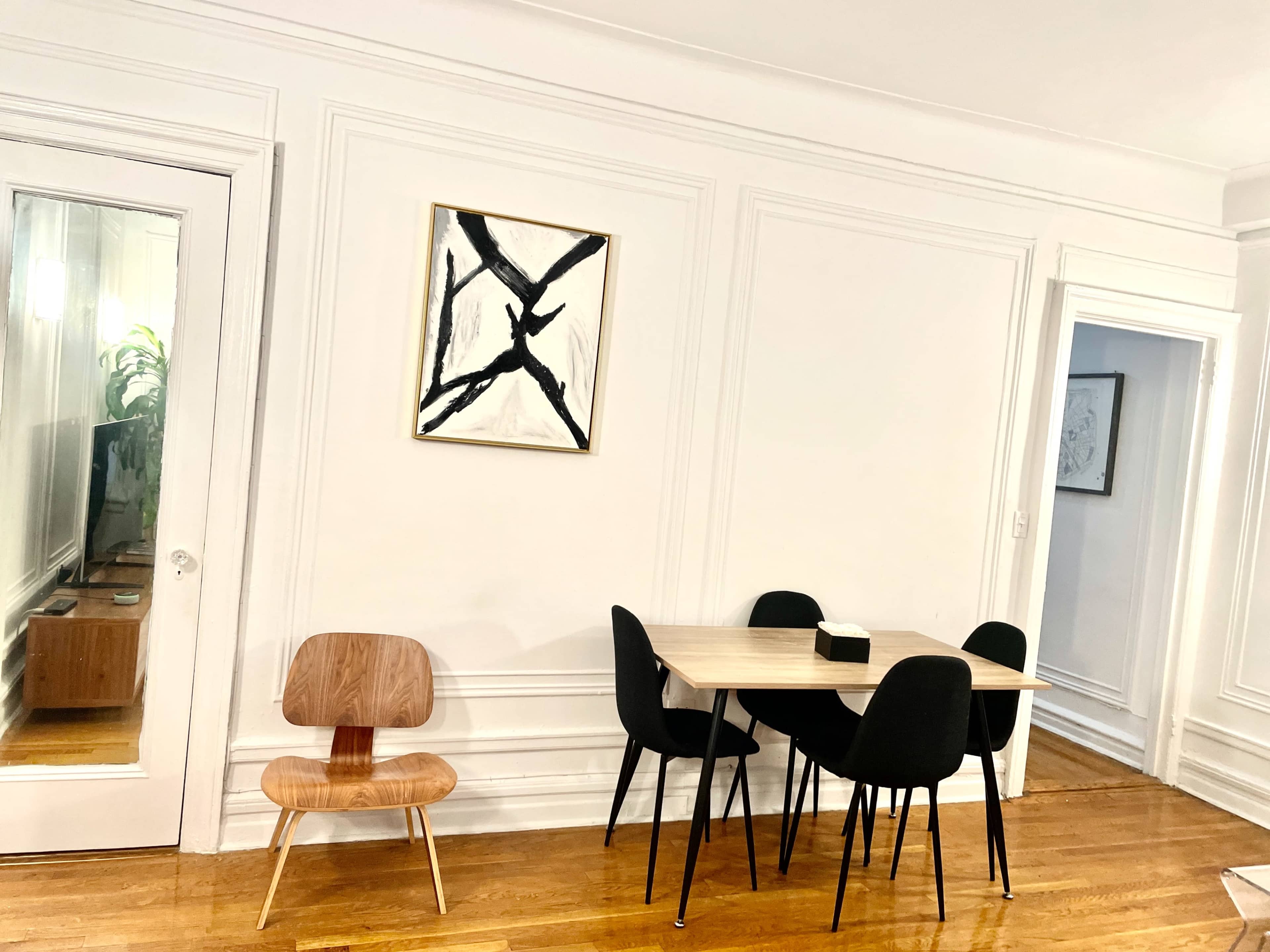 A modern dining area features a wooden table surrounded by black chairs, with a piece of abstract art hanging on the white wall.