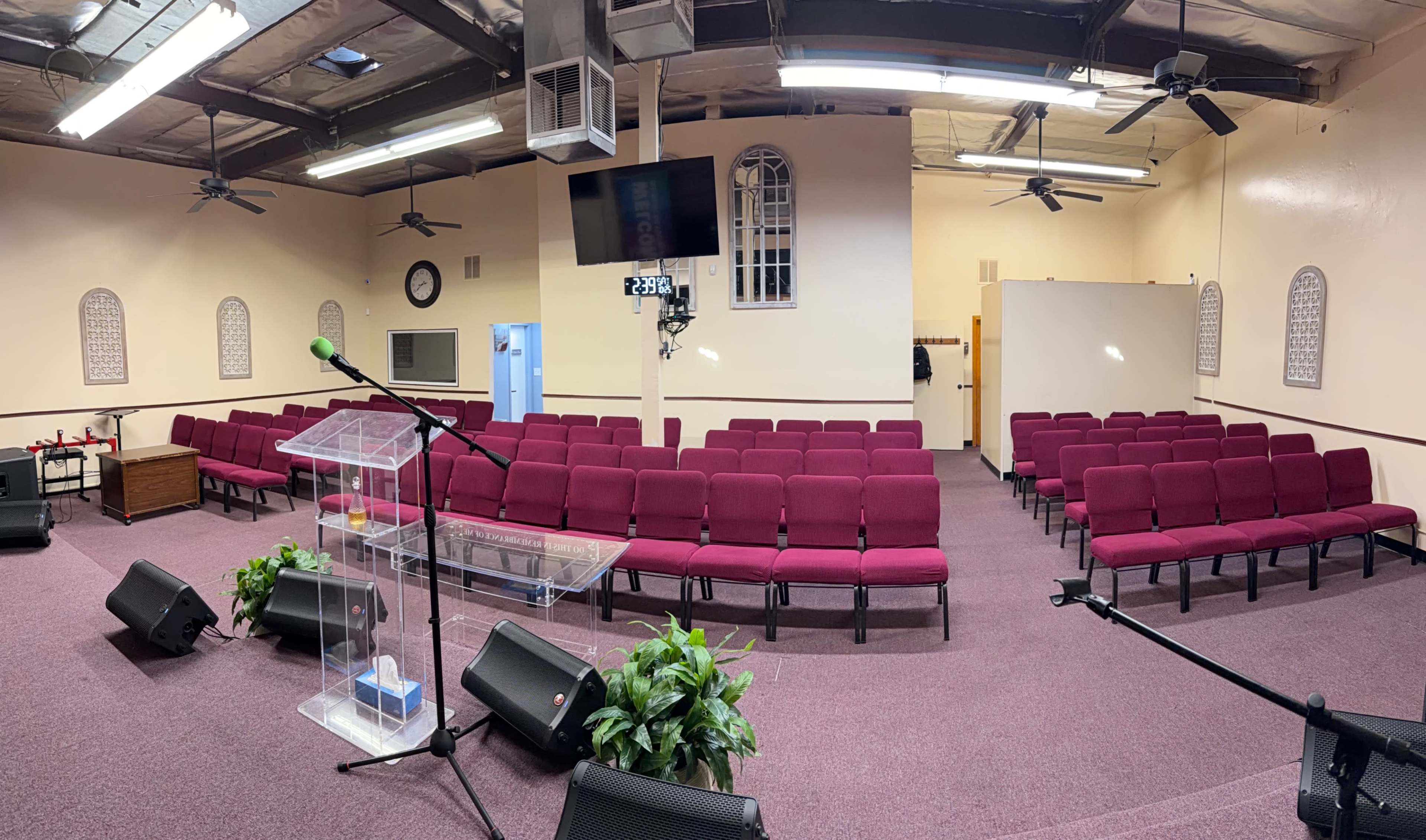The image shows a spacious, empty church interior featuring burgundy chairs arranged in rows, a transparent pulpit, and various audio equipment.