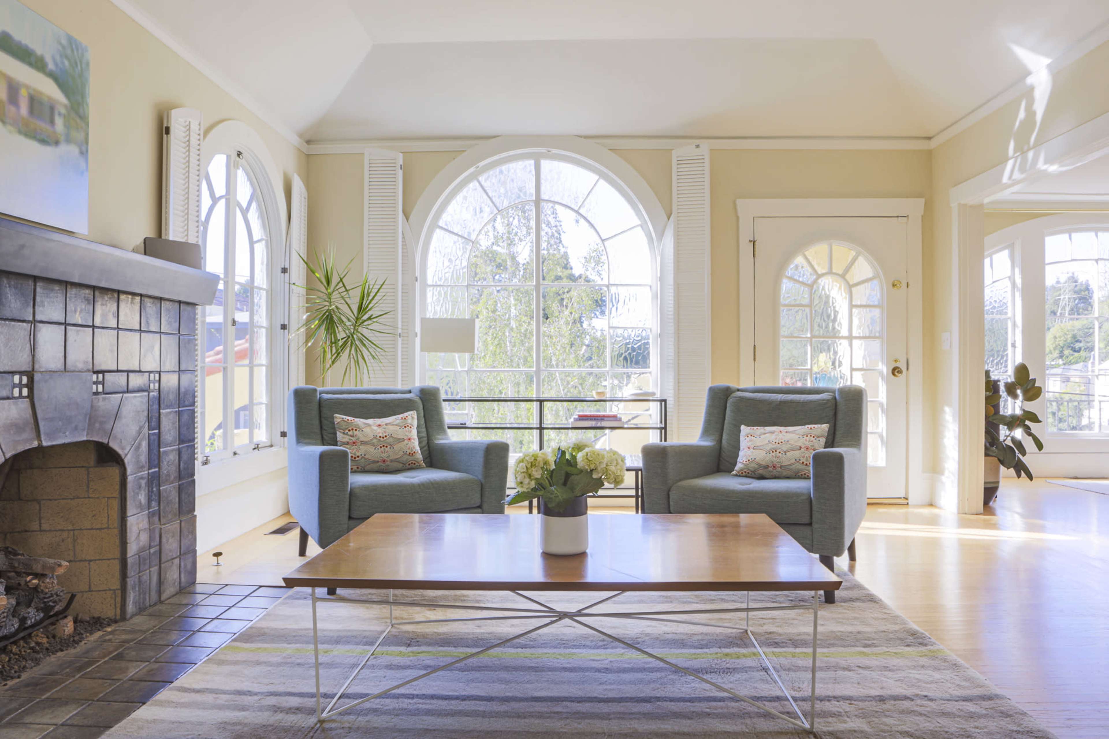 A spacious living room with two blue chairs facing a wooden coffee table, a large window letting in natural light, and a fireplace along one wall.