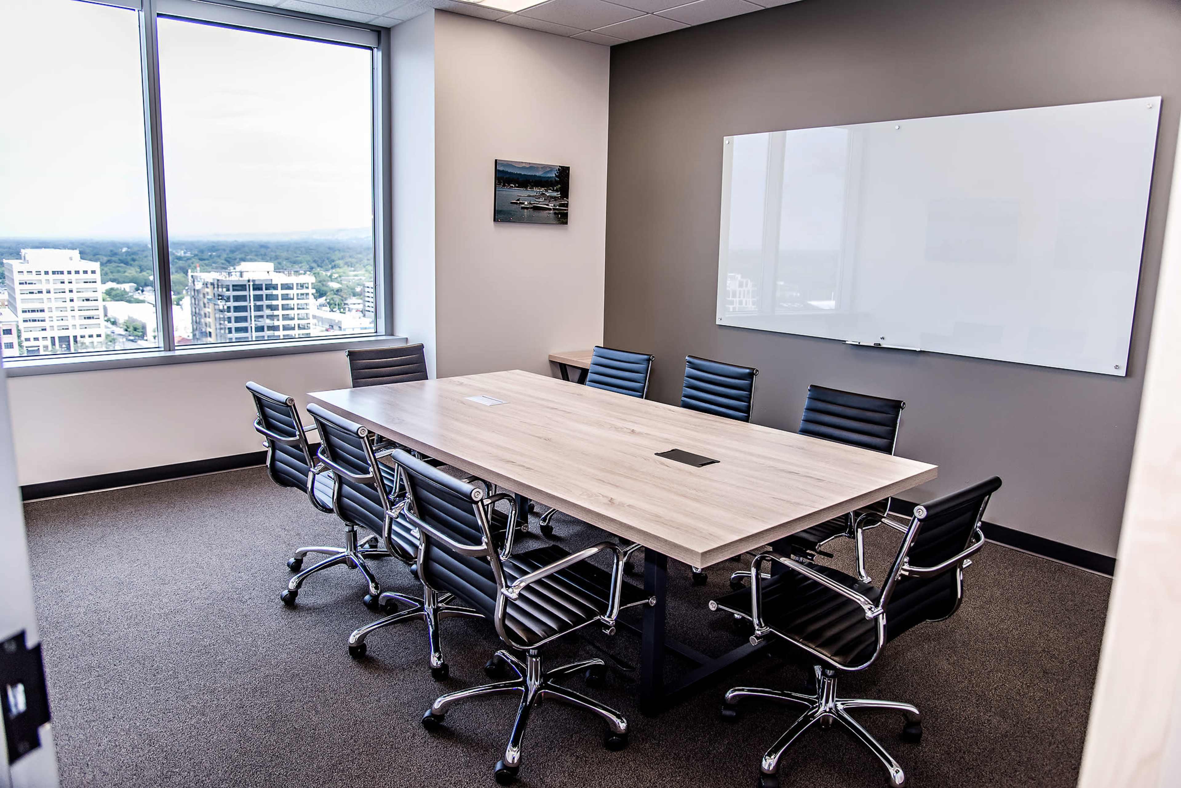 A modern conference room features a long table surrounded by black swivel chairs, with large windows offering a view of the city and a whiteboard on the wall.
