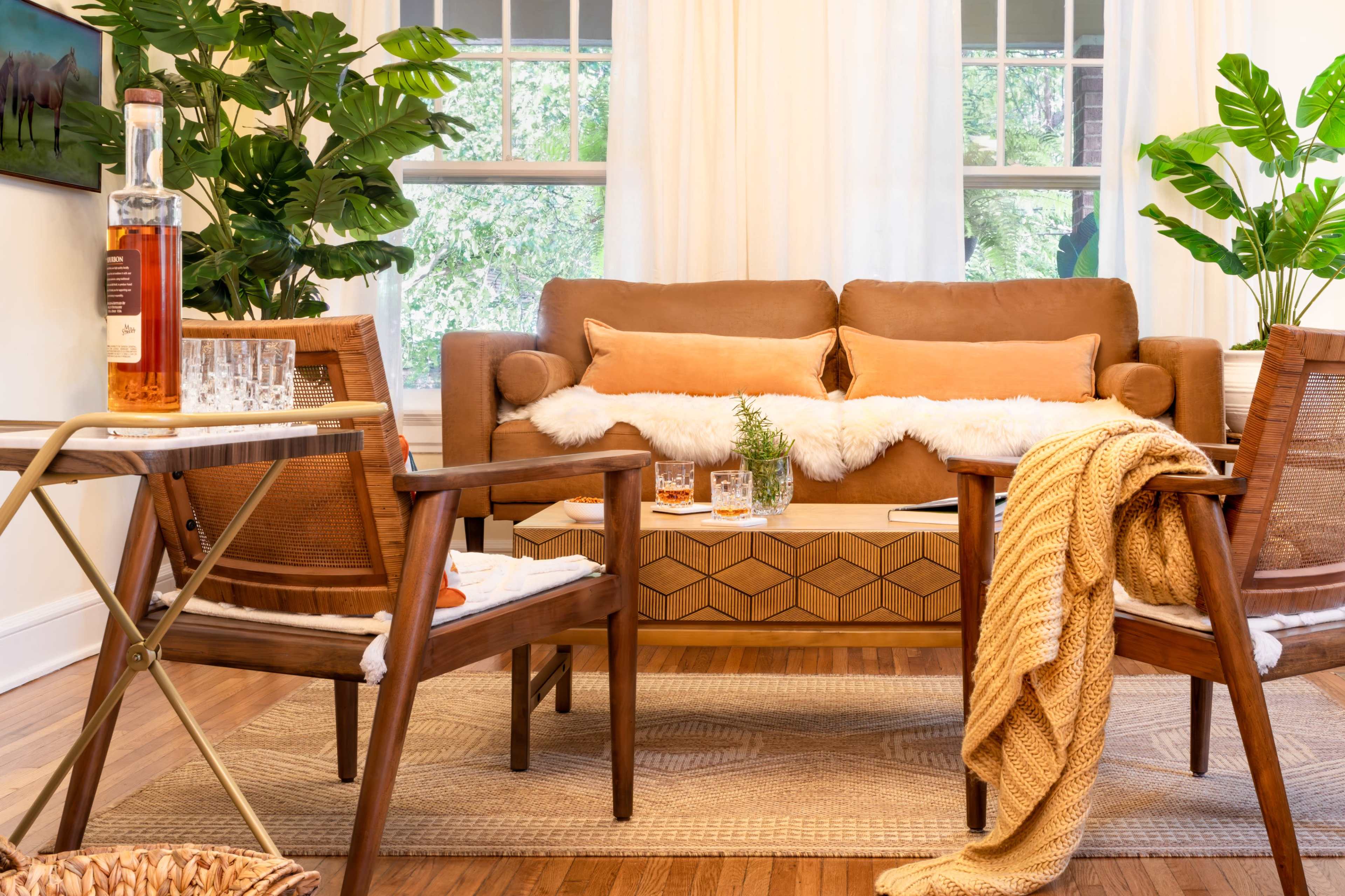 A cozy living room features a brown sofa with orange cushions, two wooden chairs, a patterned coffee table, and decorative plants, creating an inviting atmosphere.
