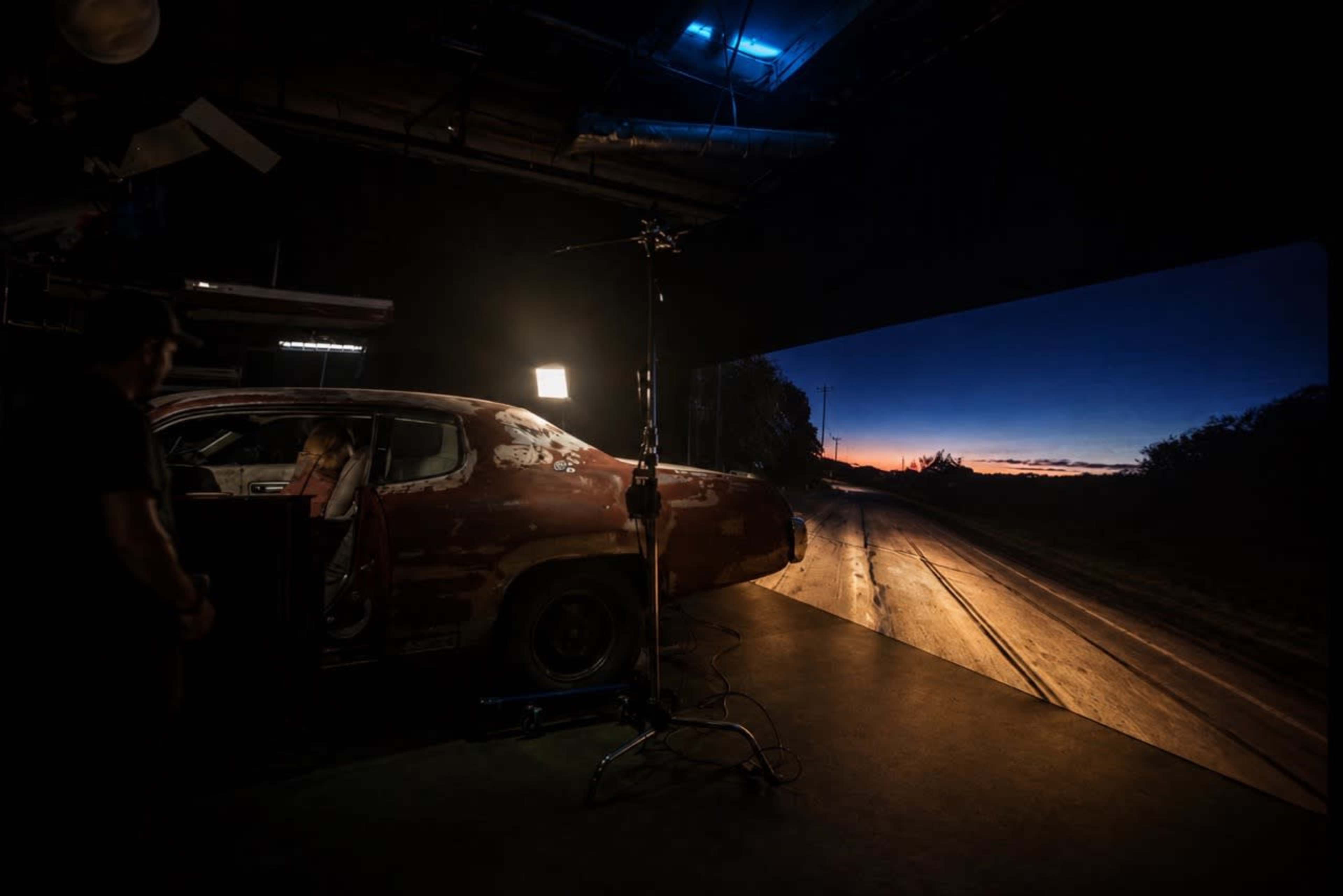 A vintage car is positioned in a studio setting, with a large backdrop displaying a twilight landscape of a road at sunset.