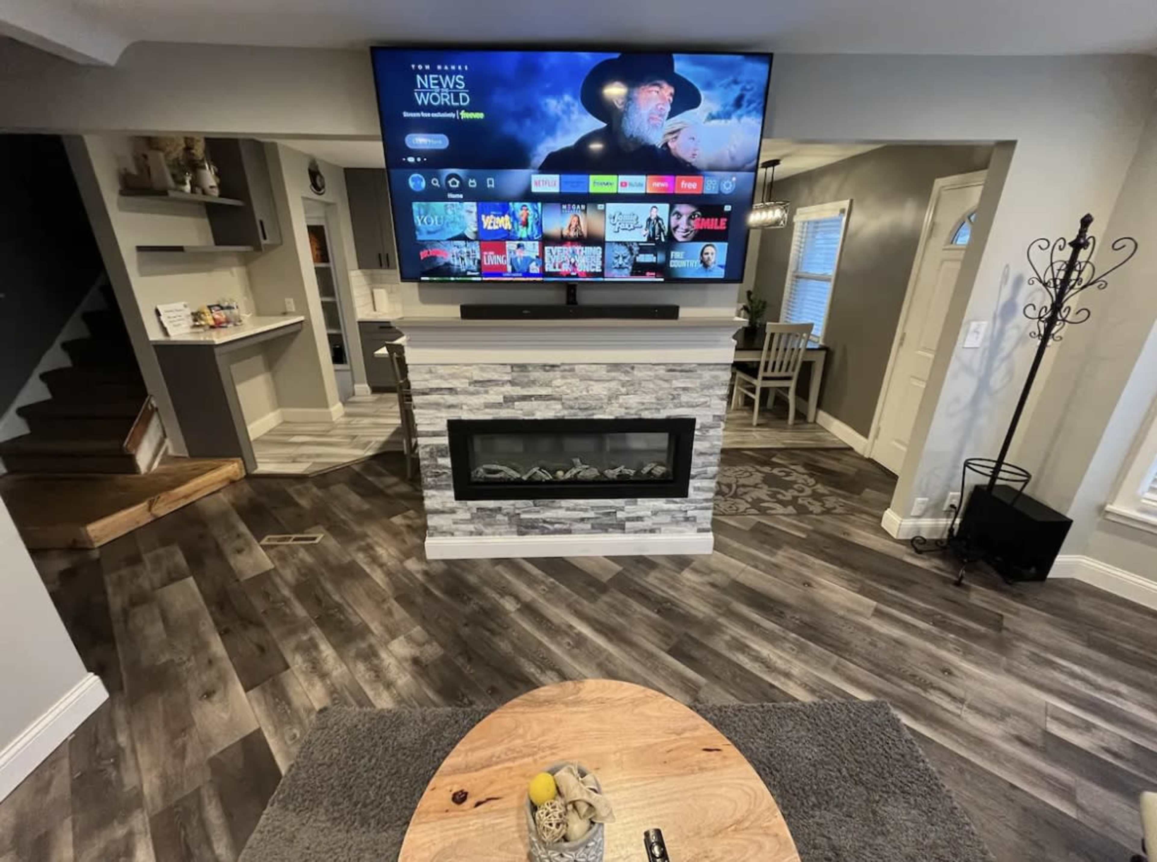 The room features a modern living area with a wall-mounted television above a stone fireplace, a wooden coffee table in the center, and a staircase in the background.