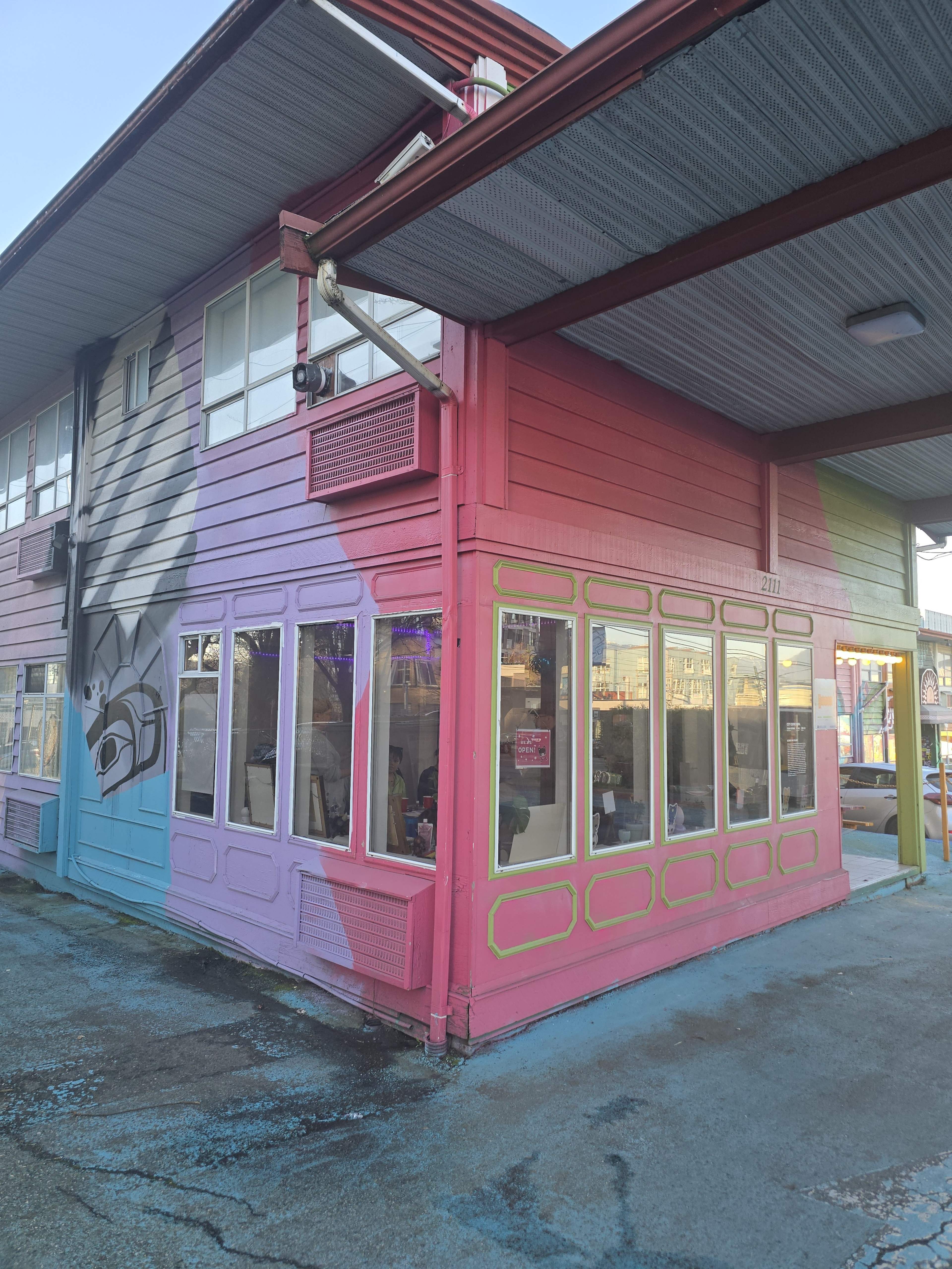The image shows a two-story building with a vibrant exterior featuring pink and blue paint, large windows, and a mural on one side.