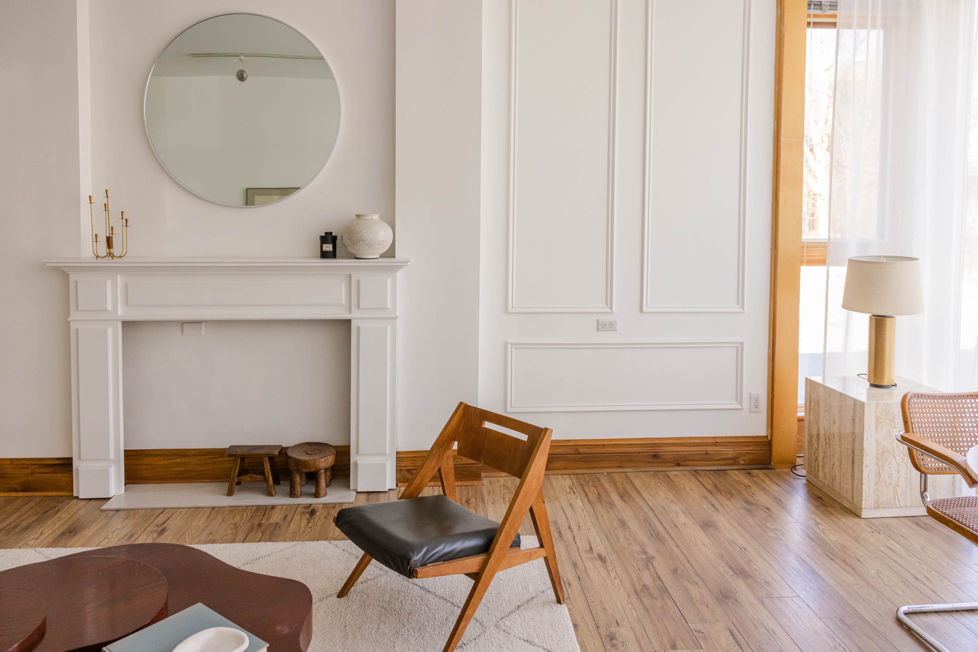 A minimalist living room features a round mirror above a white mantel, a wooden chair, and natural light coming through a window.