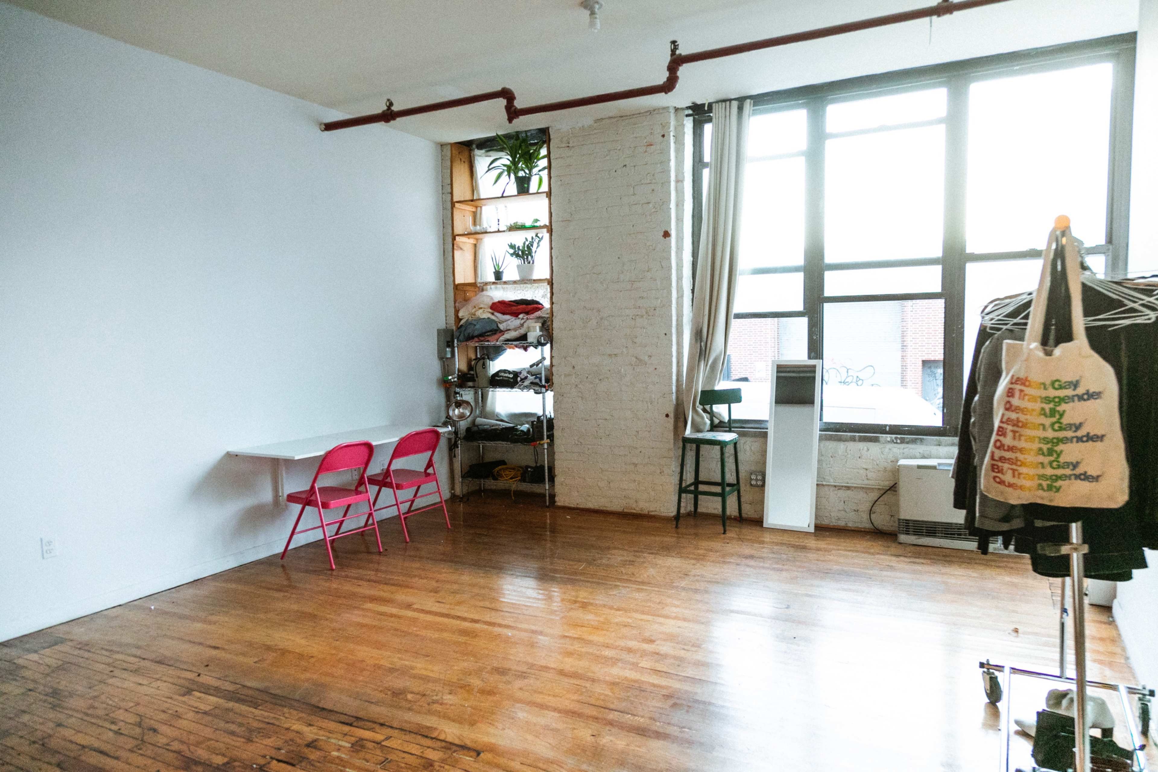 The spacious room features hardwood floors, a large window, a small table with two pink chairs, and a shelving unit stacked with clothes.