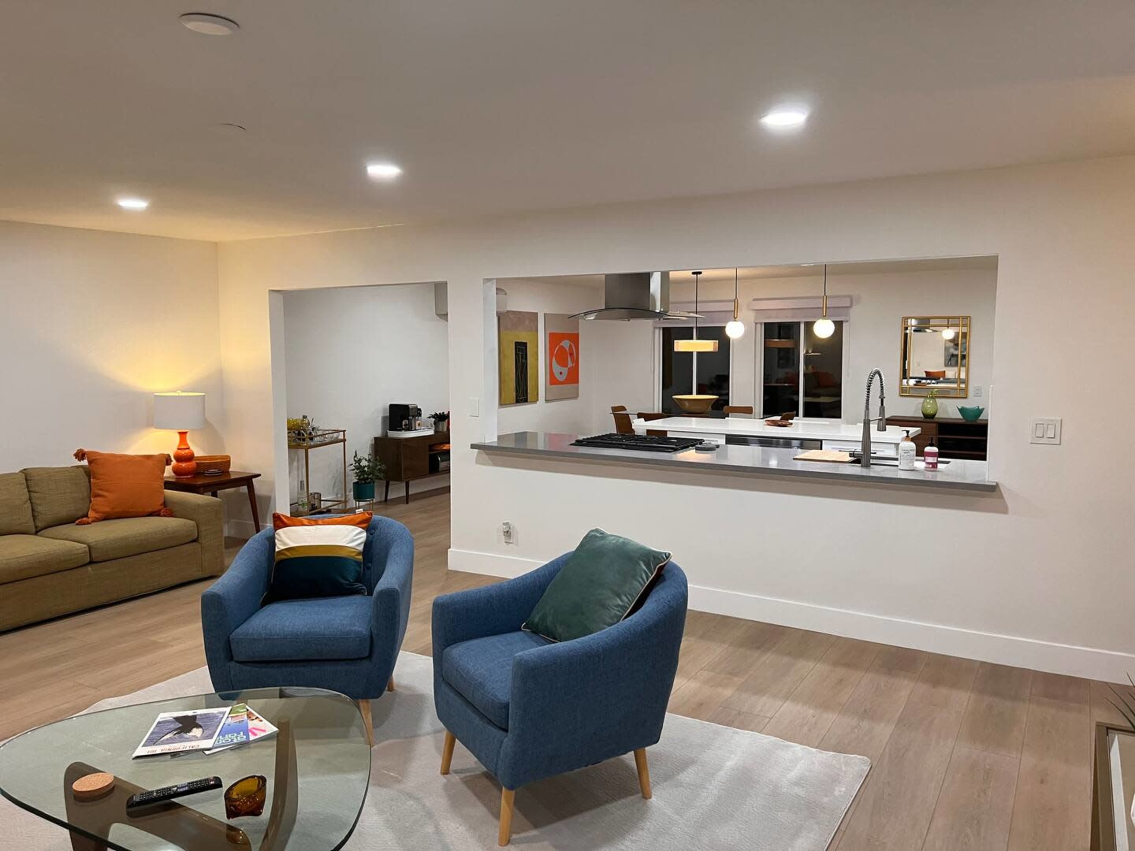 A modern living space featuring a green sofa, two blue chairs, a glass coffee table, and an open kitchen area with a bar counter and pendant lights.