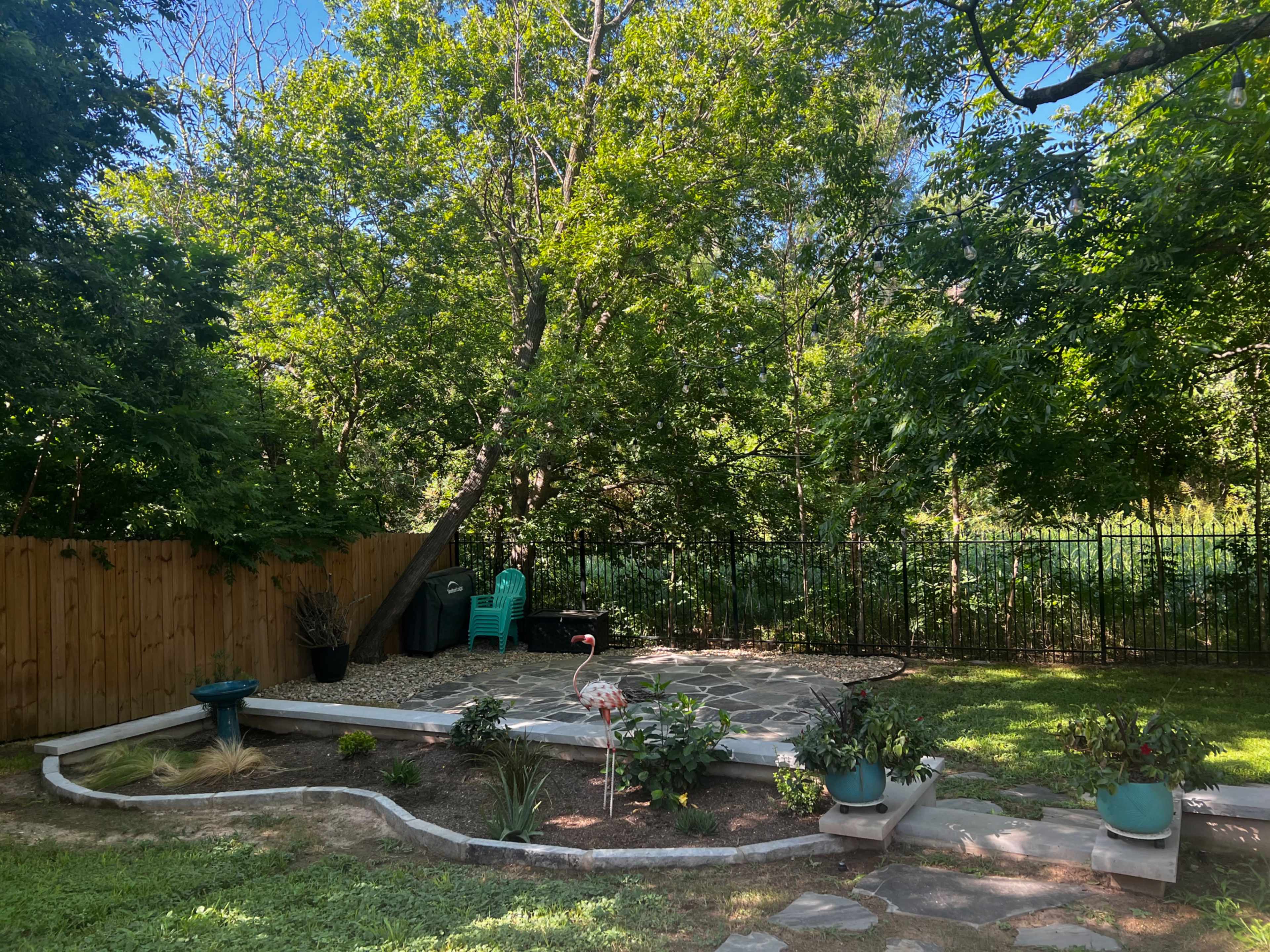A backyard garden with a stone patio, potted plants, and a fence surrounding a wooded area.
