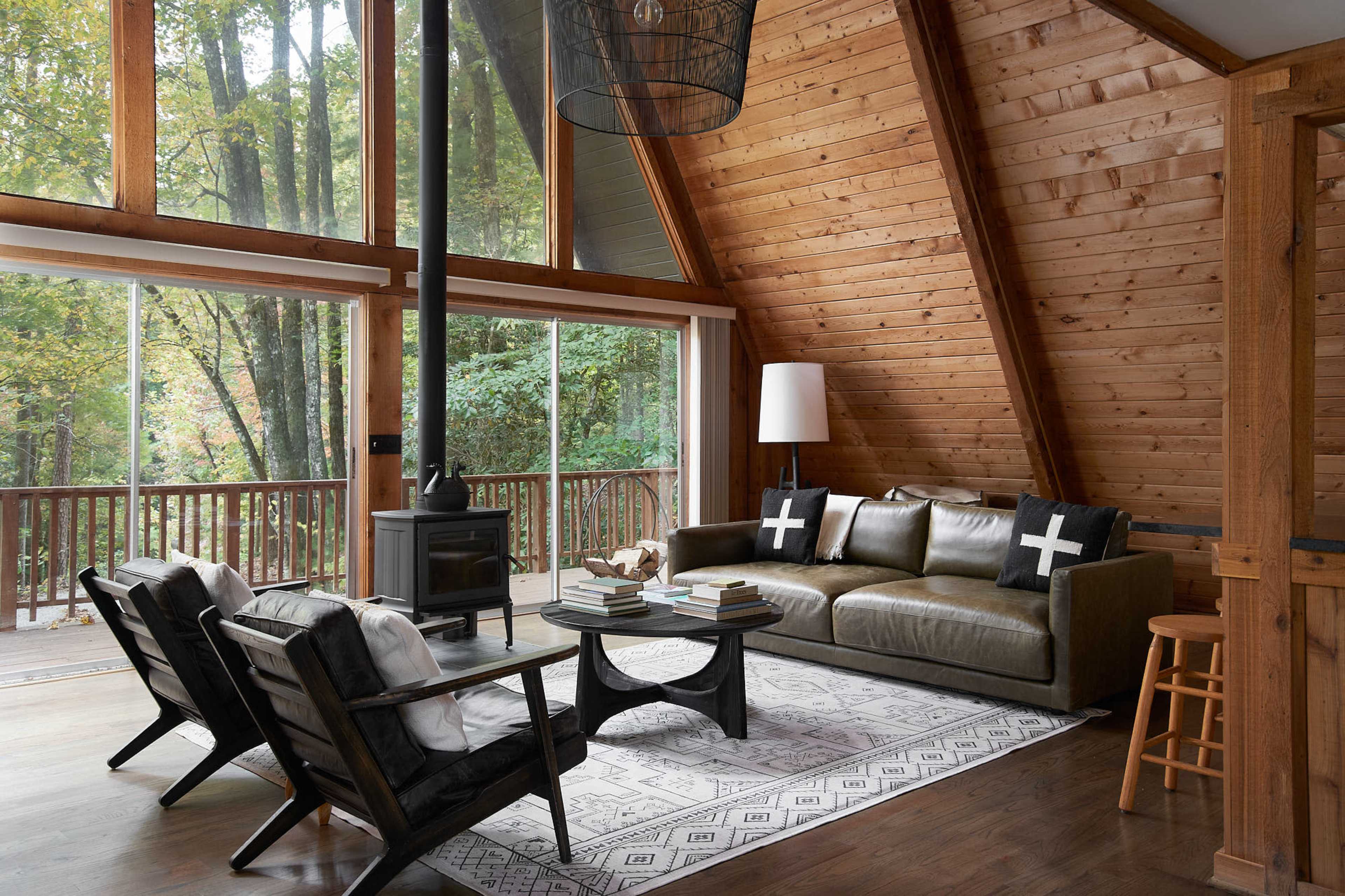 A wooden A-frame living room features a sofa, two black chairs, a circular coffee table, and large windows overlooking a forested area.