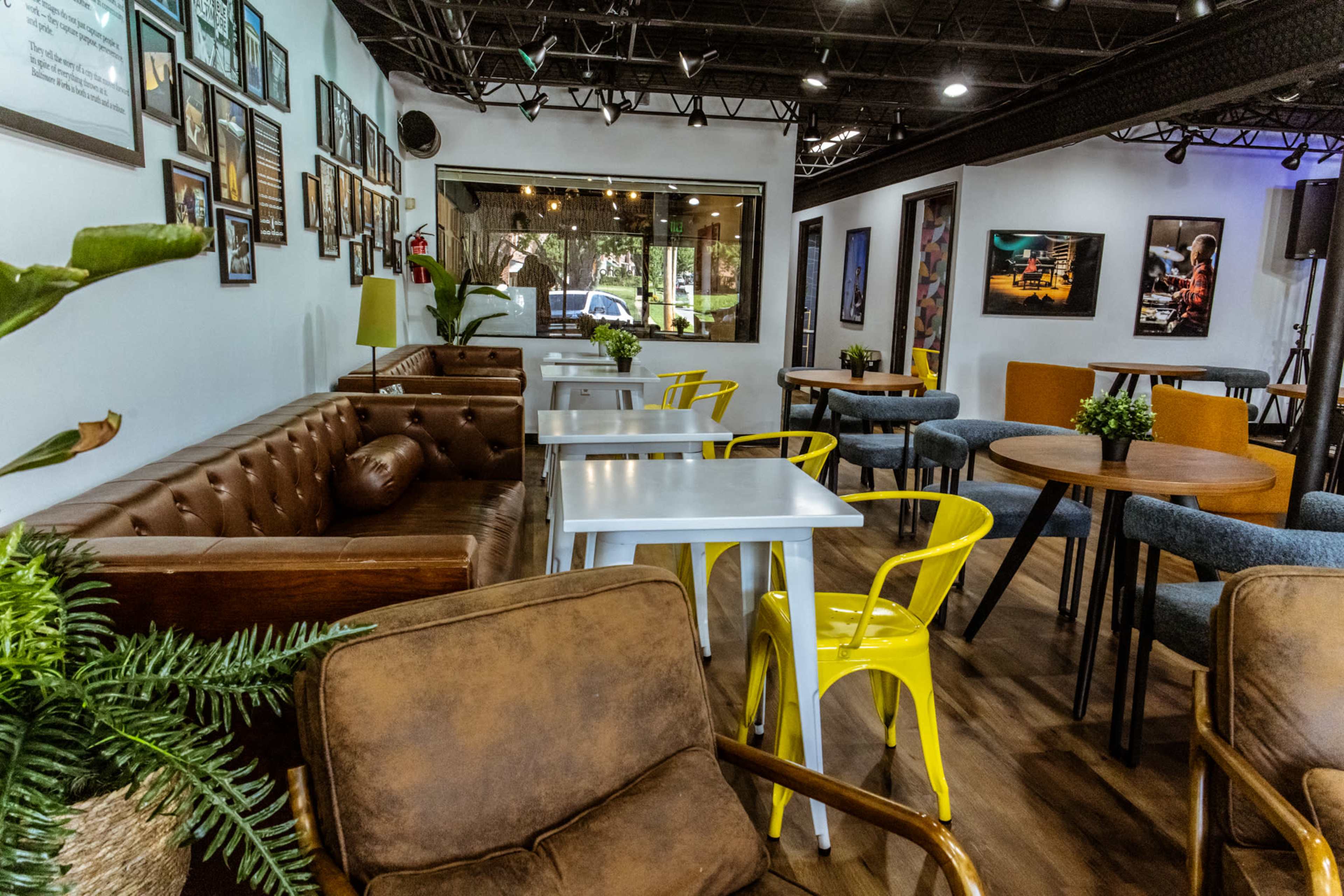 The image shows a modern café interior featuring a mix of brown leather and yellow chairs around white and wooden tables, with framed photographs on the walls.