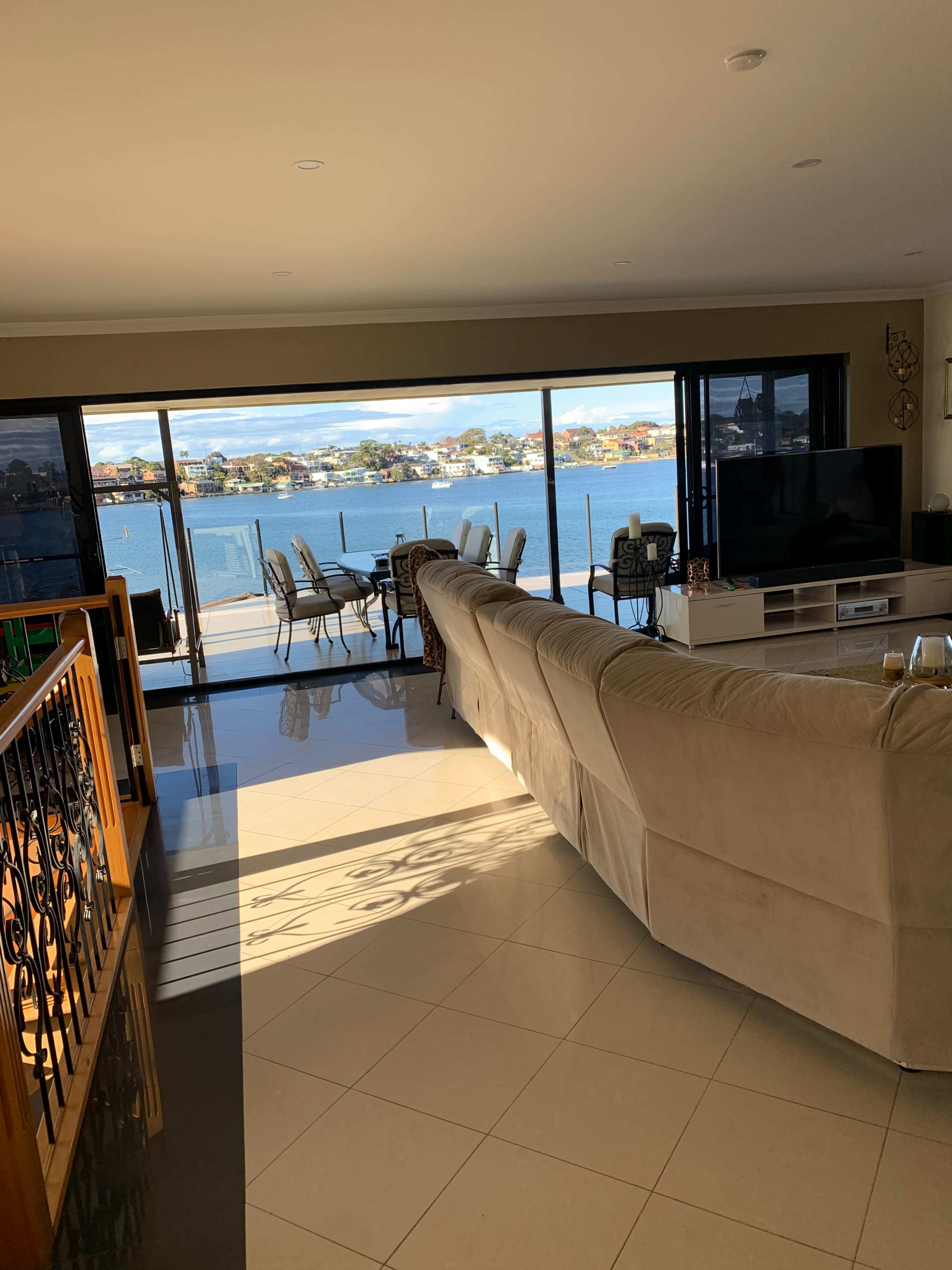 A spacious living room with large glass doors opens to a balcony overlooking a waterway, featuring a beige sectional sofa and a television.