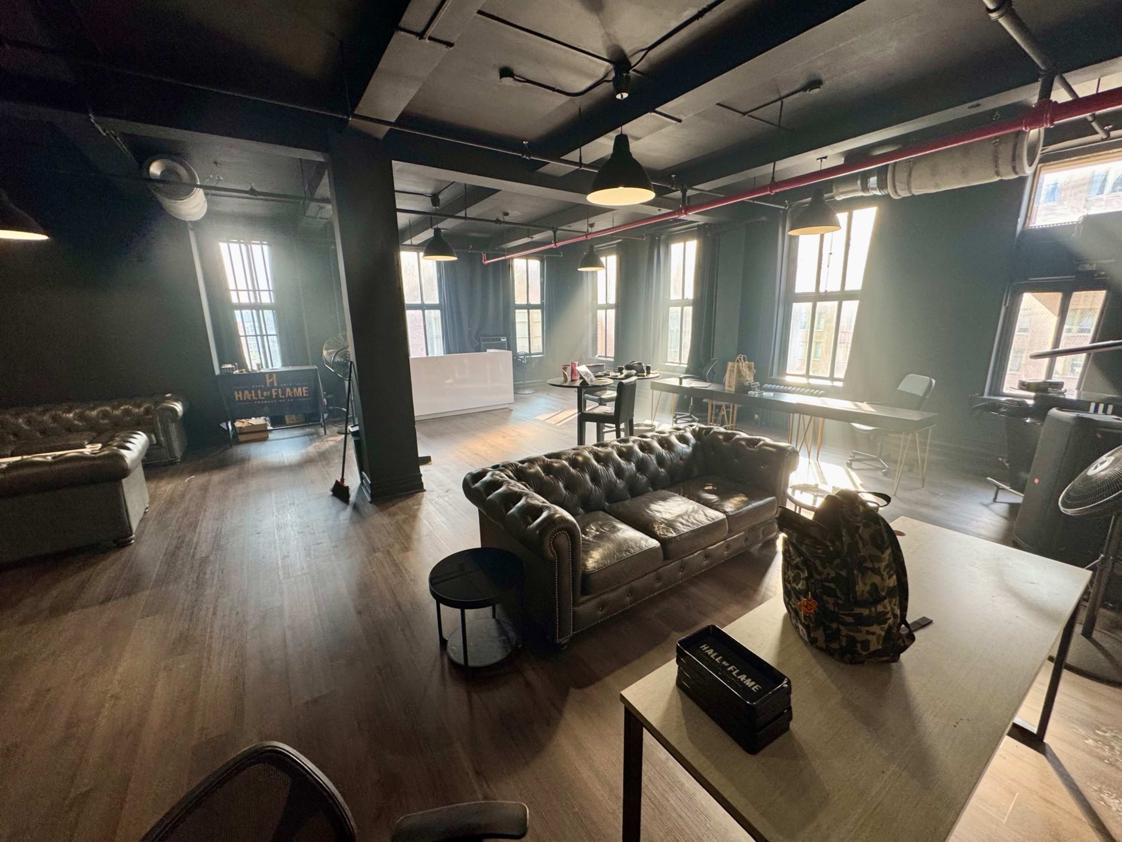 The image shows a spacious, modern room with dark walls, multiple windows, and a mix of seating arrangements, including a leather sofa and tables.