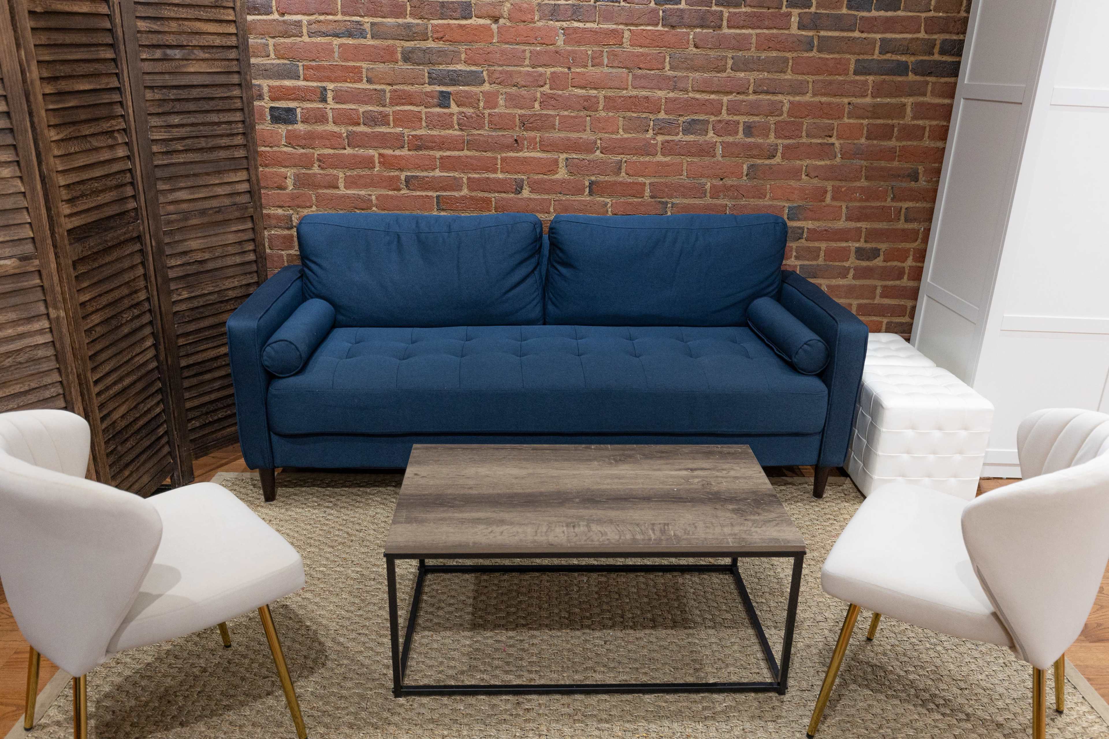 A blue couch is positioned in front of a wooden coffee table on a woven rug, surrounded by two white chairs against a brick wall.