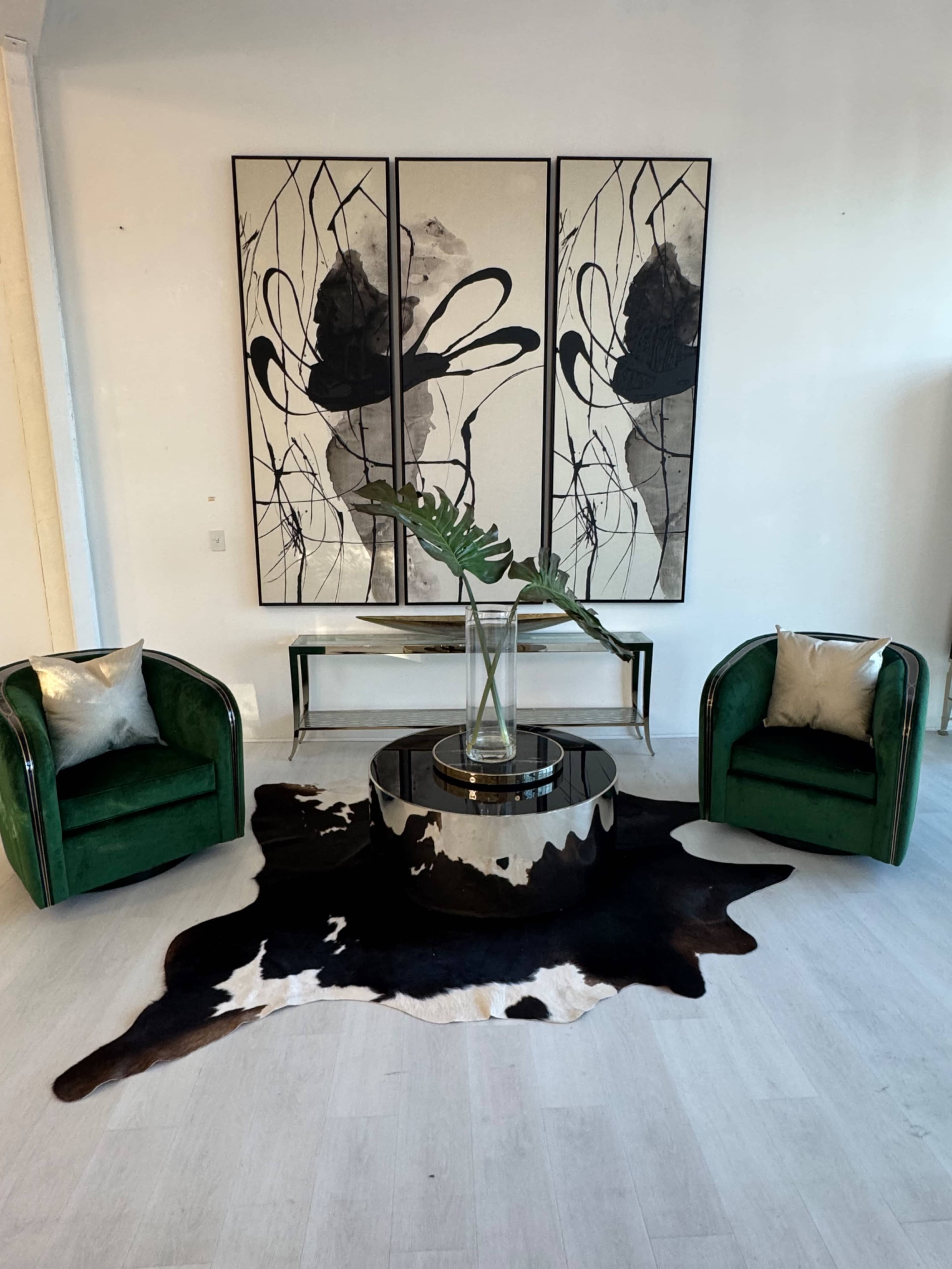 The image shows a stylish interior with two green chairs flanking a round coffee table, set on a cowhide rug beneath artwork featuring abstract figures on the wall.