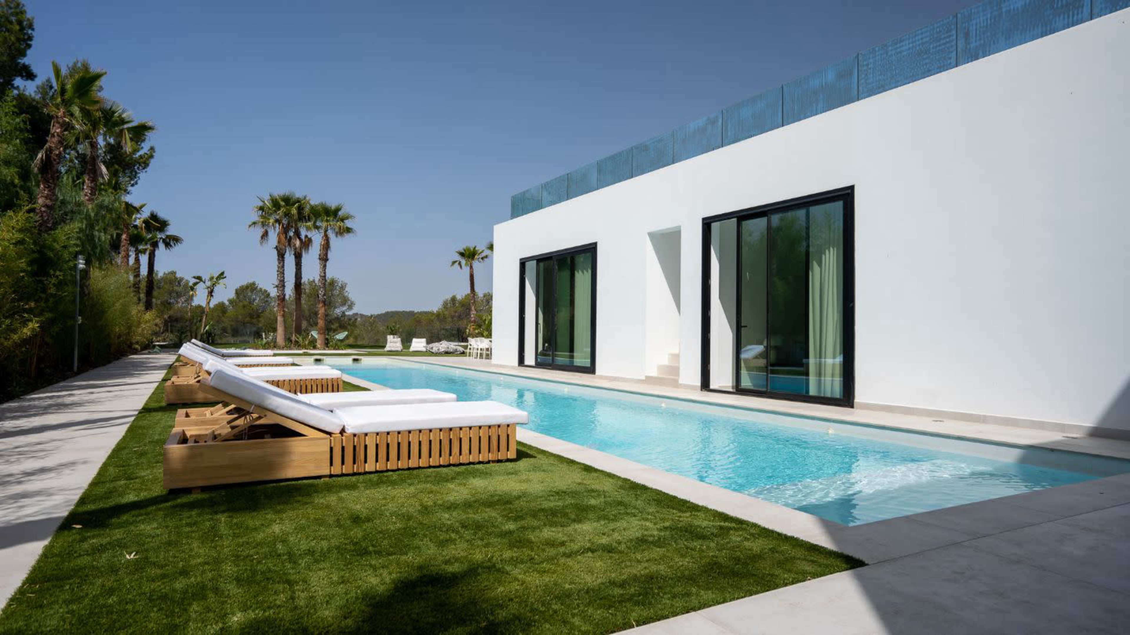 A modern villa features a rectangular swimming pool bordered by palm trees and lounge chairs on a grassy area.