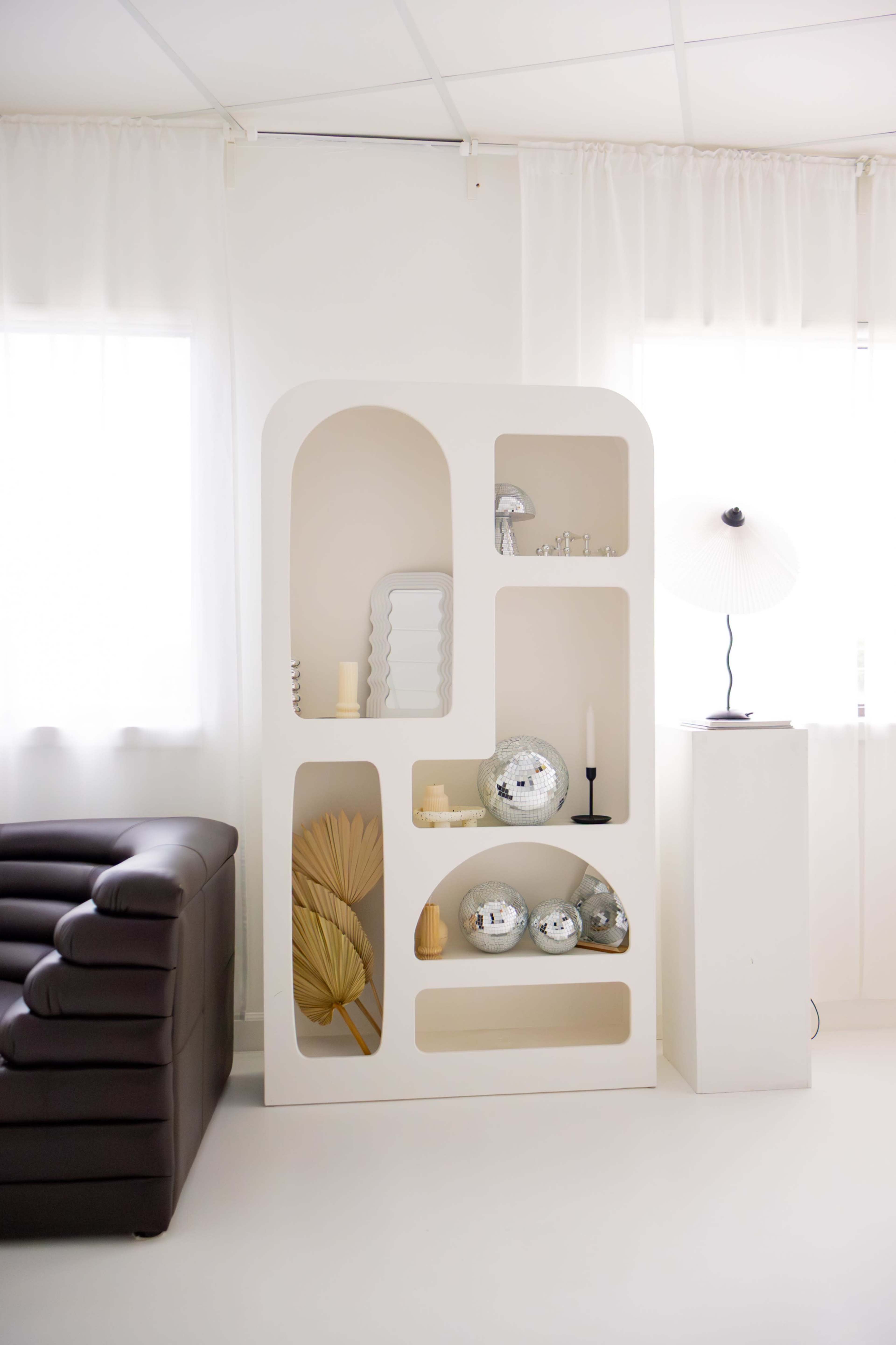 A modern white shelving unit with decorative items is positioned in a bright room next to a brown leather couch.