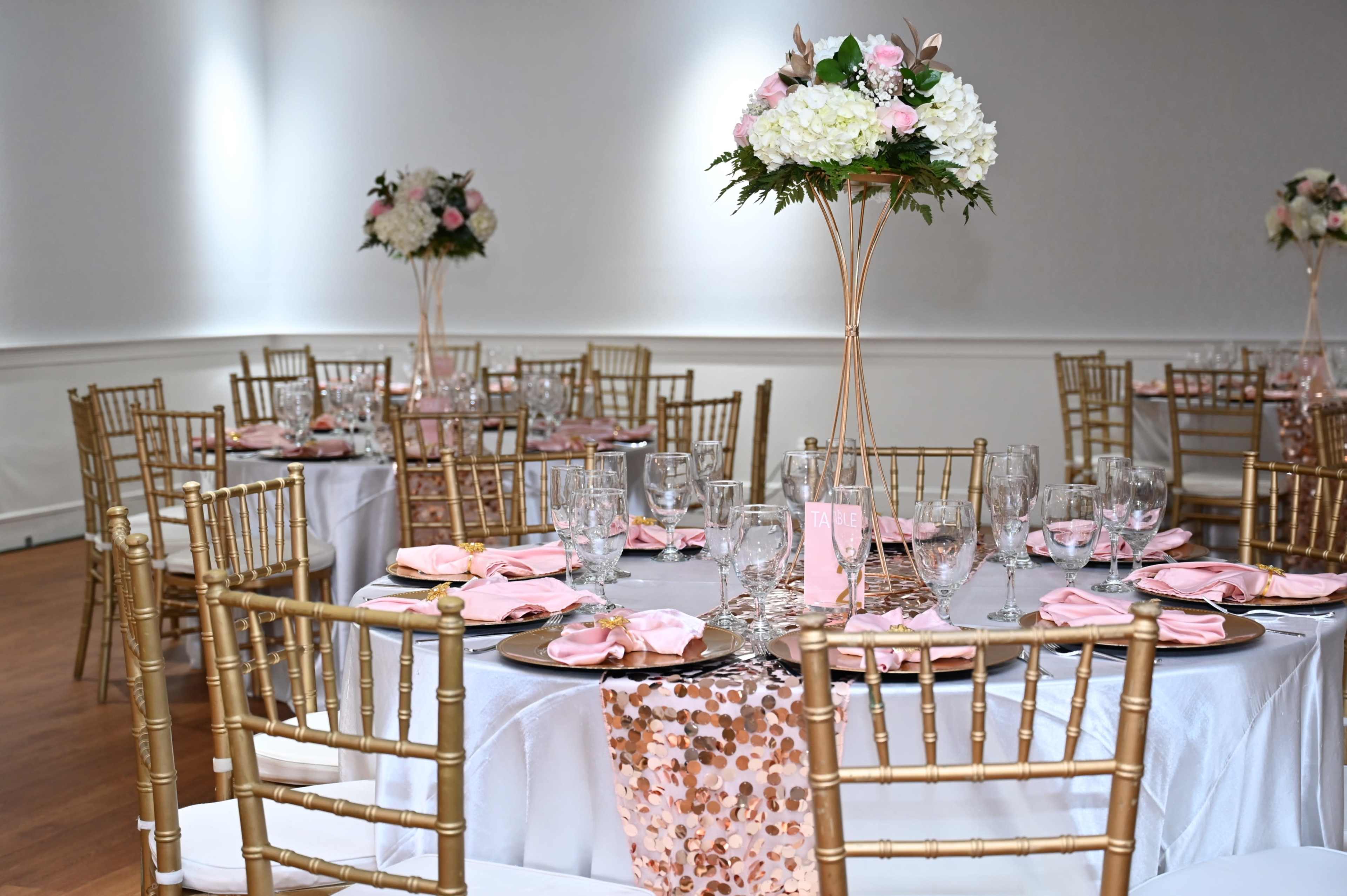 A banquet room is set with elegantly arranged tables featuring gold chiavari chairs, pink napkins, and floral centerpieces.