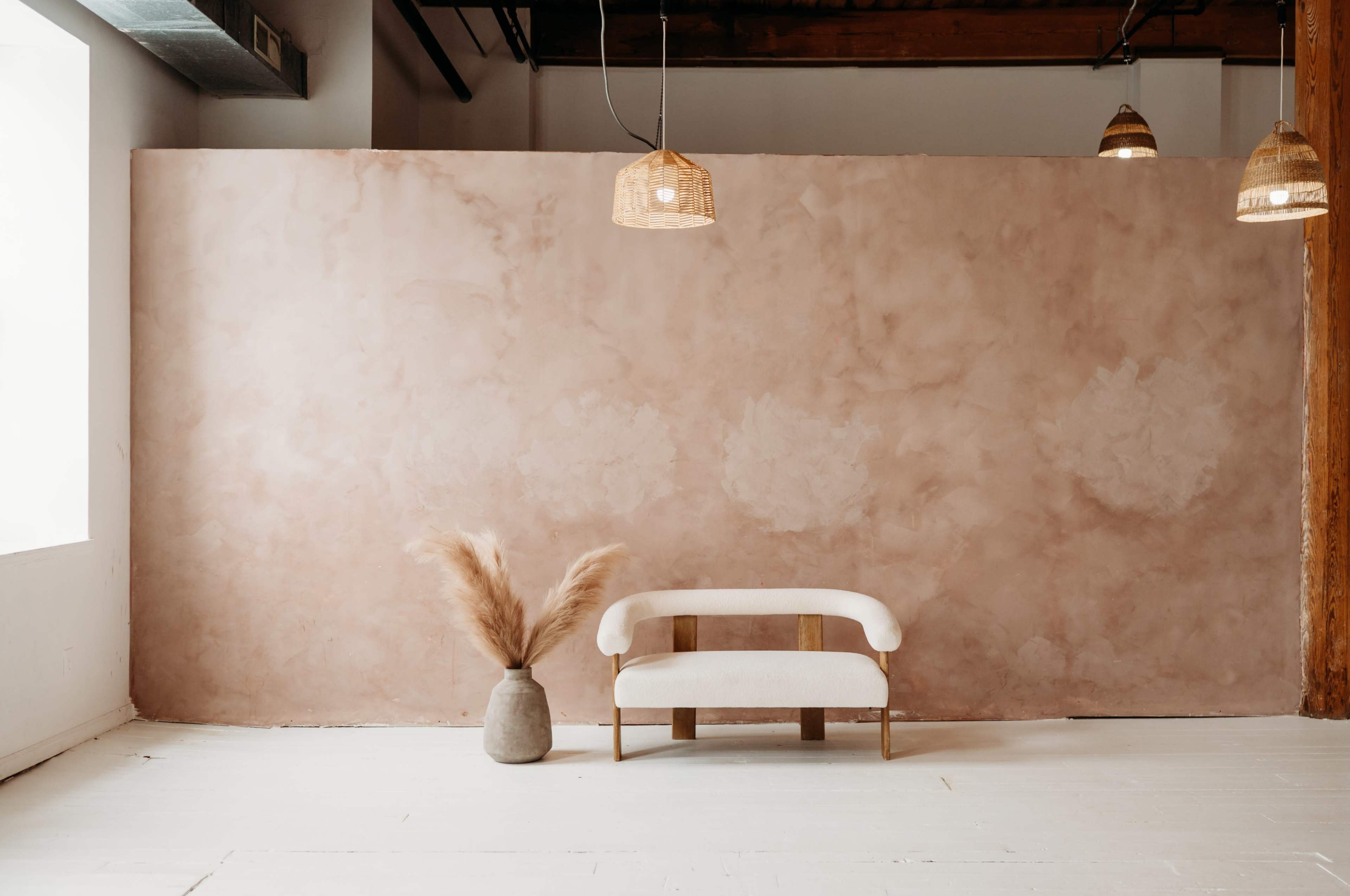 A minimalist interior features a pale pink textured wall, a white sofa with gold accents, and a pampas grass arrangement in a vase.