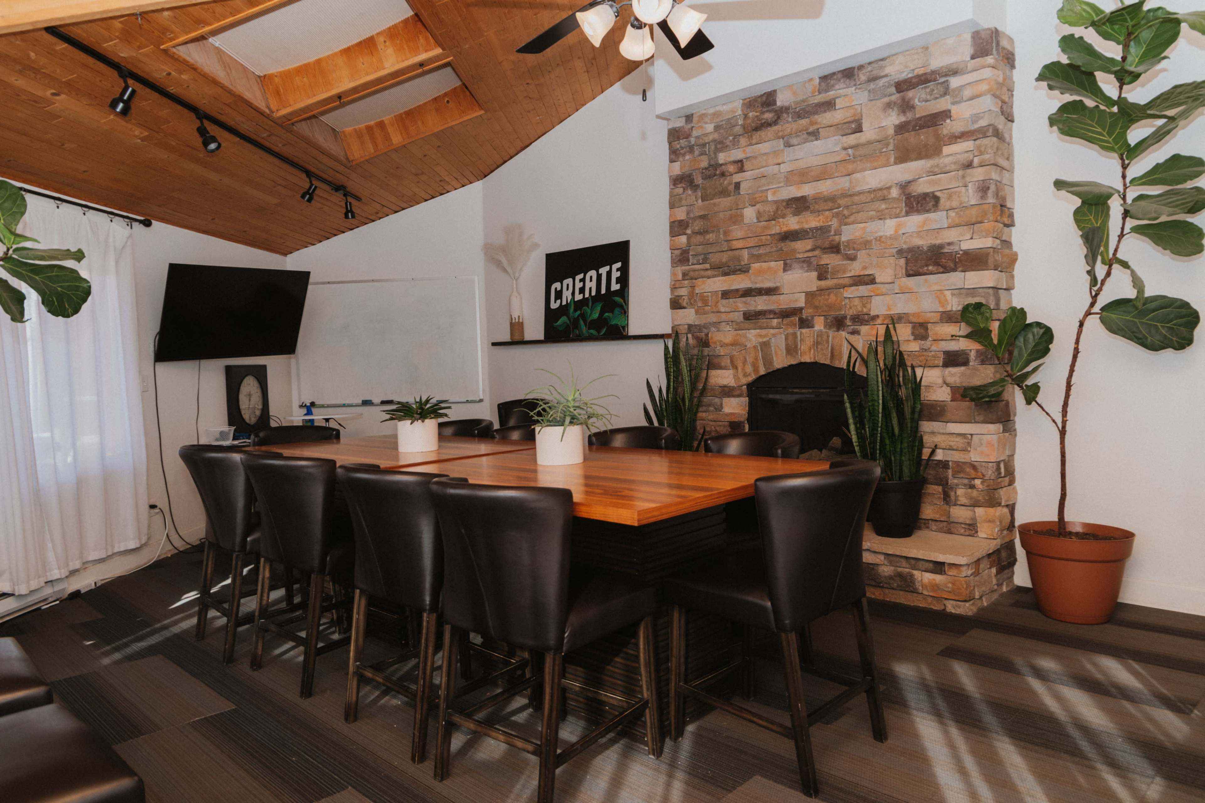 The image shows a modern conference room with a large wooden table surrounded by black chairs, a stone accent wall, a television, and potted plants.