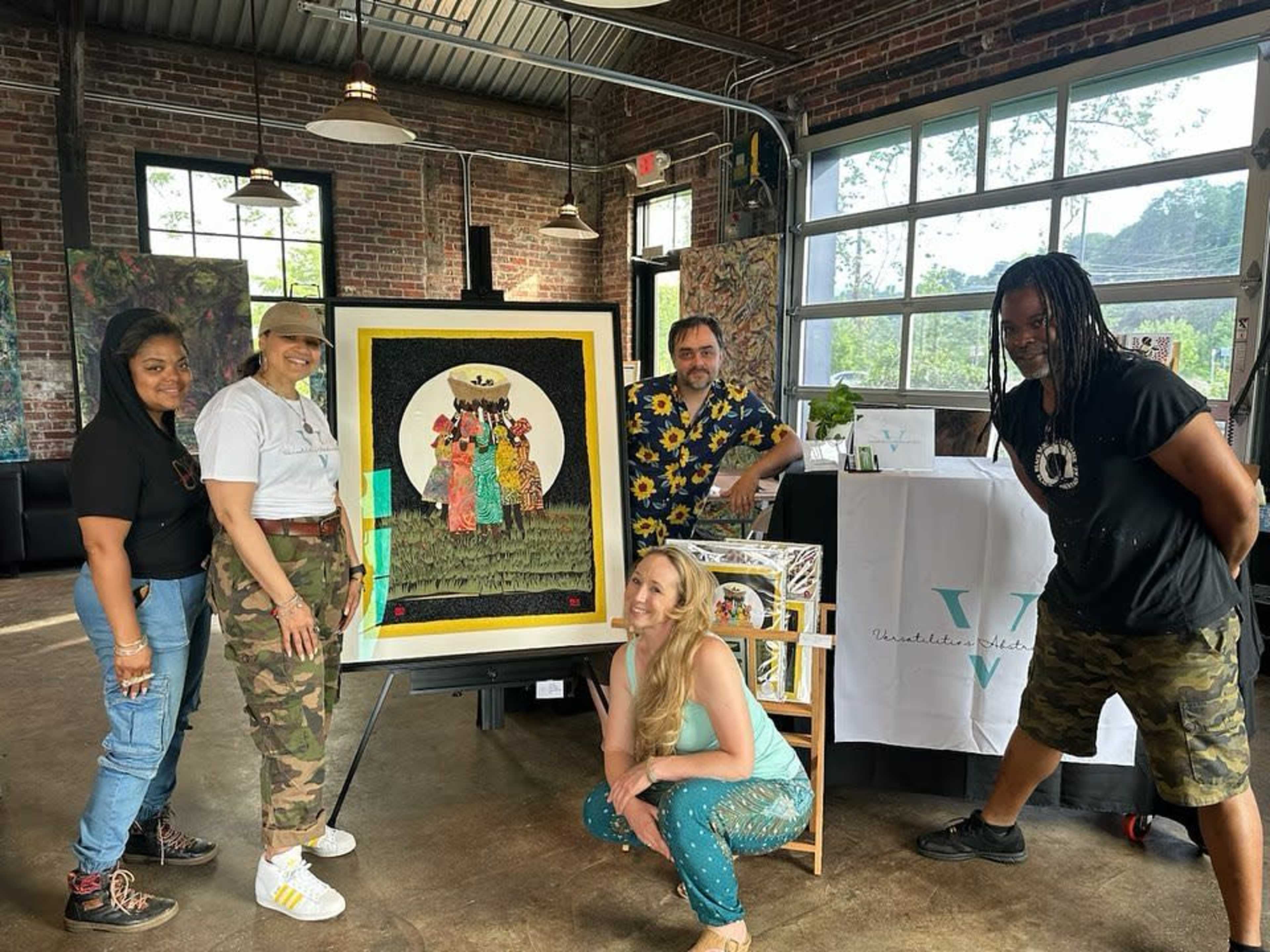 A group of four individuals poses around a large framed artwork in a modern, industrial-style venue with exposed brick walls and large windows.