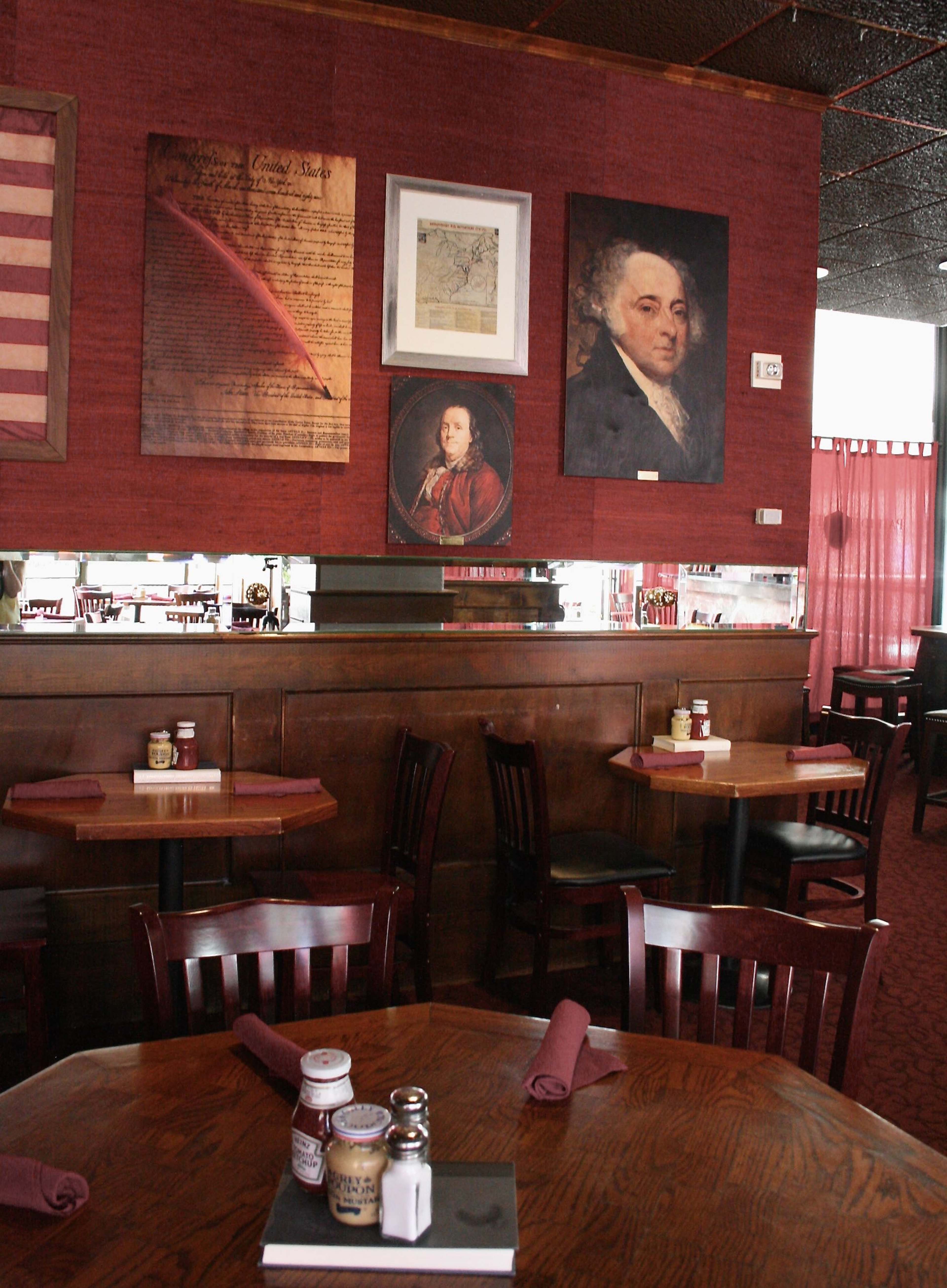 A restaurant interior featuring dark wood furniture, red walls adorned with historical artwork, and table settings with condiments and napkins.