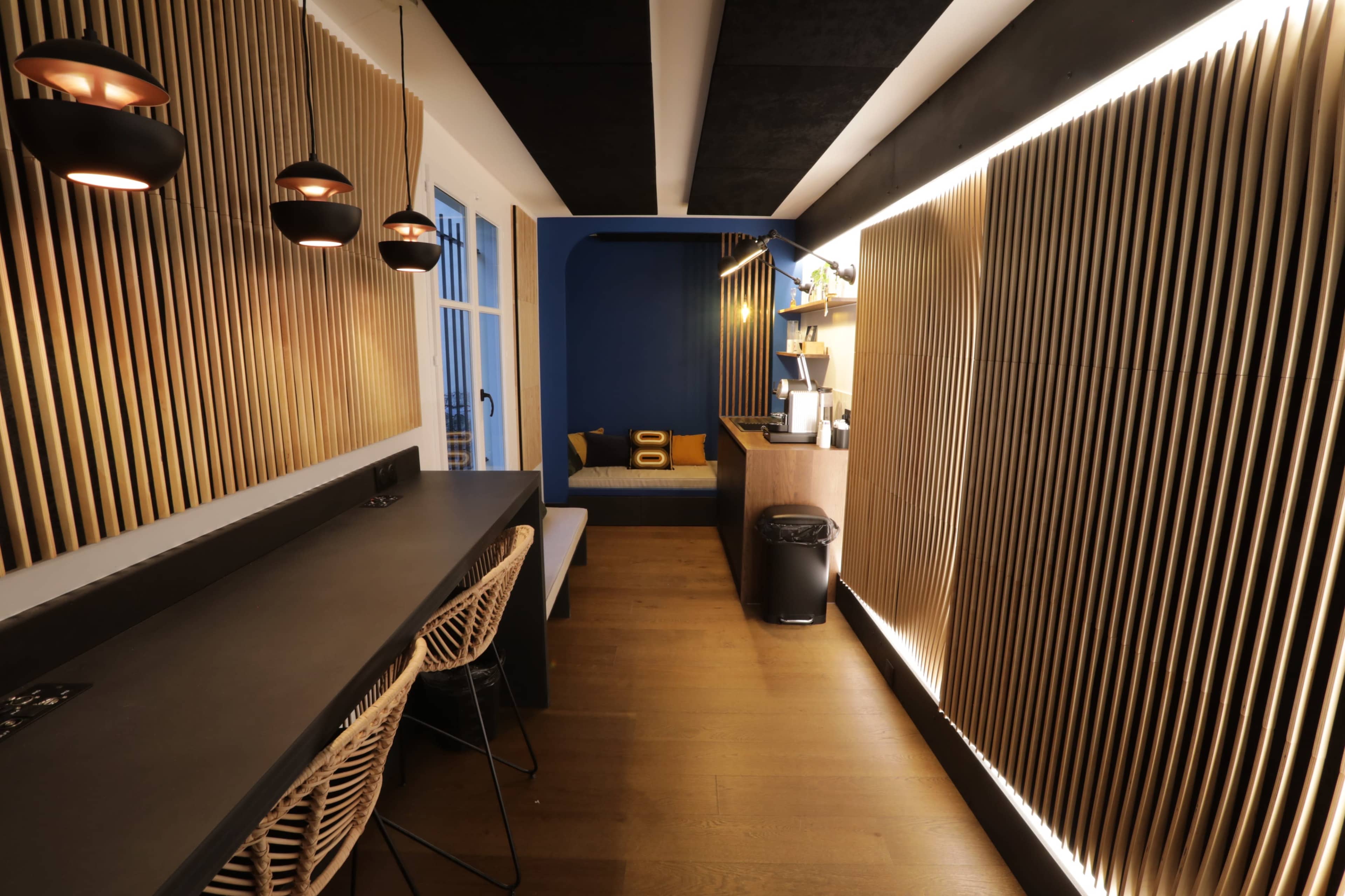 A modern interior space featuring a long black countertop with bar stools, wooden slatted walls, and a cozy seating area illuminated by pendant lights.