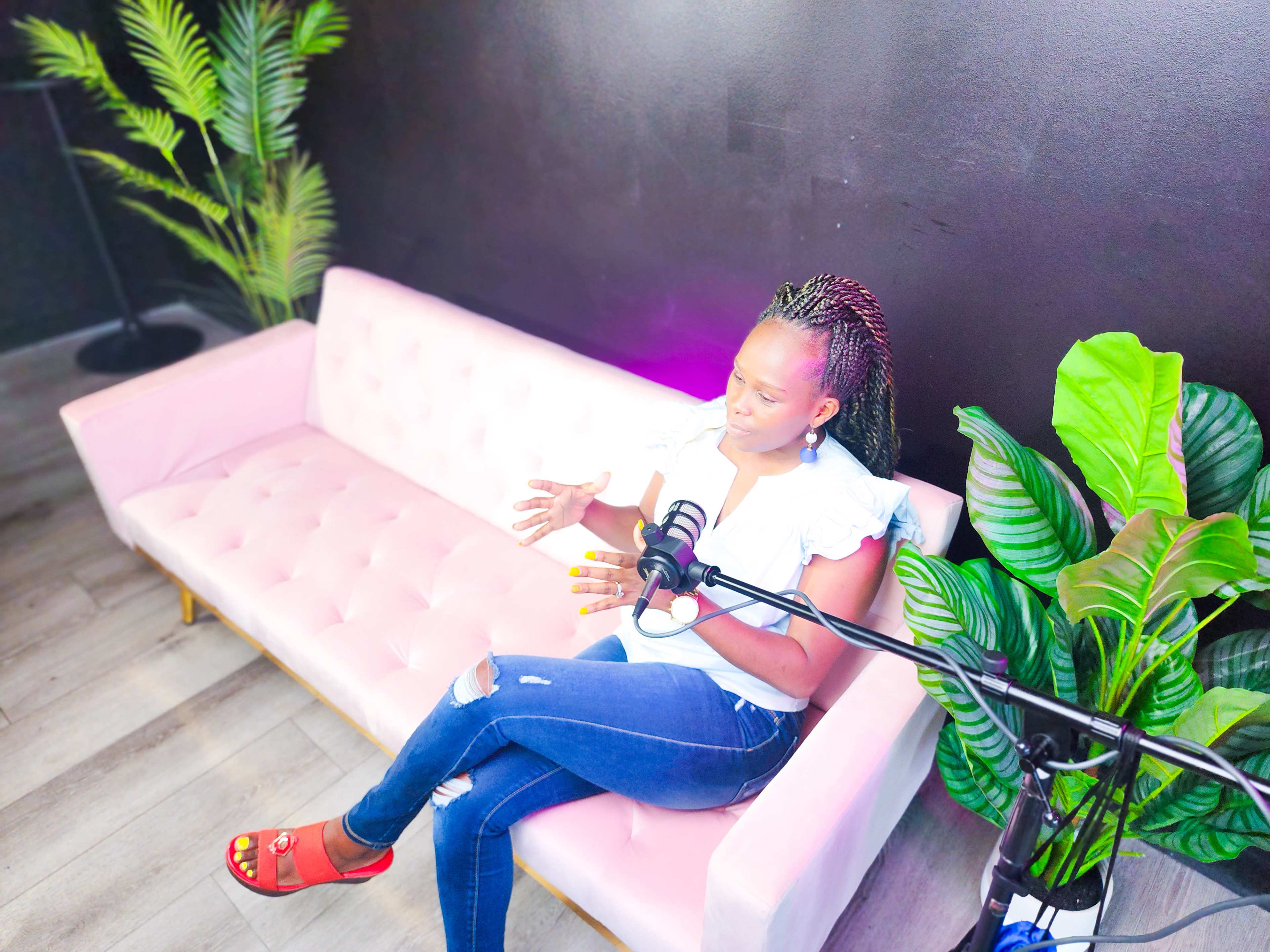 A woman is sitting on a pink sofa, talking into a microphone, with potted plants in the background.