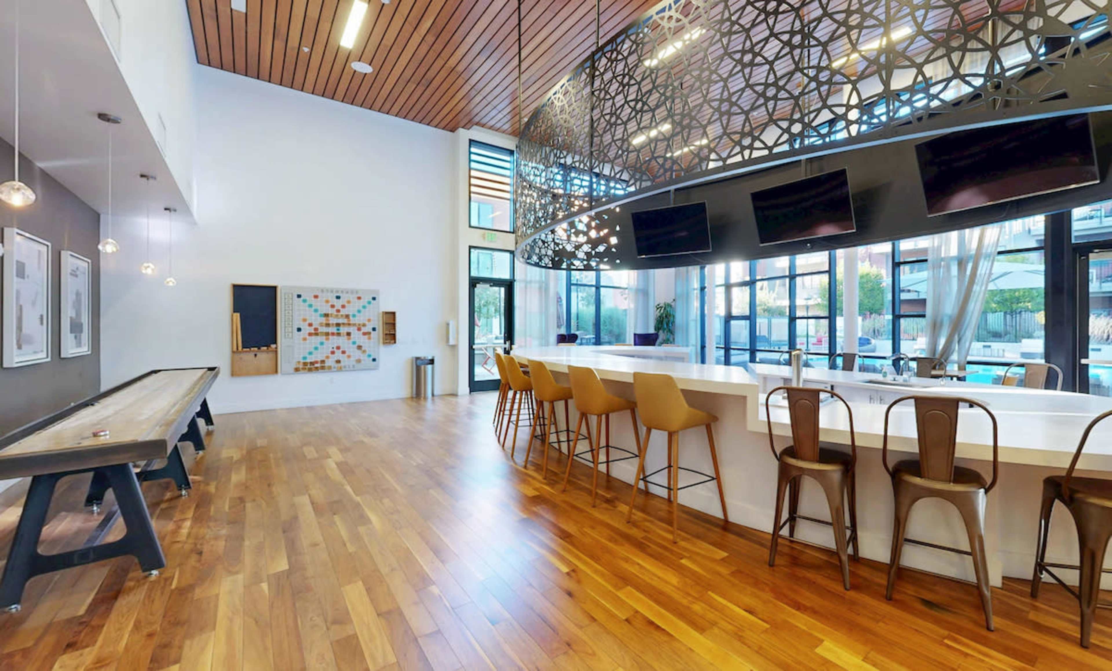 The image shows a modern common area with a bar, seating, a table for games, large windows, and a decorative light fixture overhead.