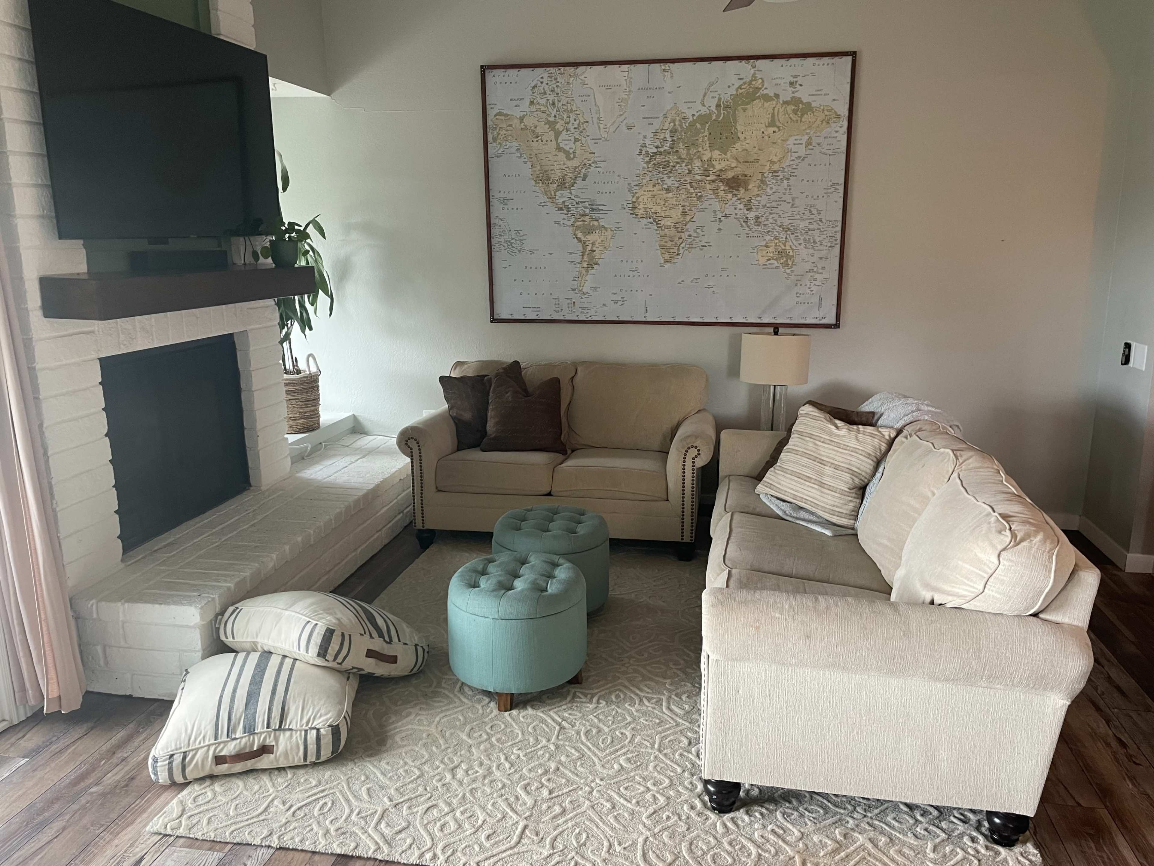 A cozy living room features a beige sofa, an armchair, a blue ottoman, and a large world map hanging on the wall above a fireplace.