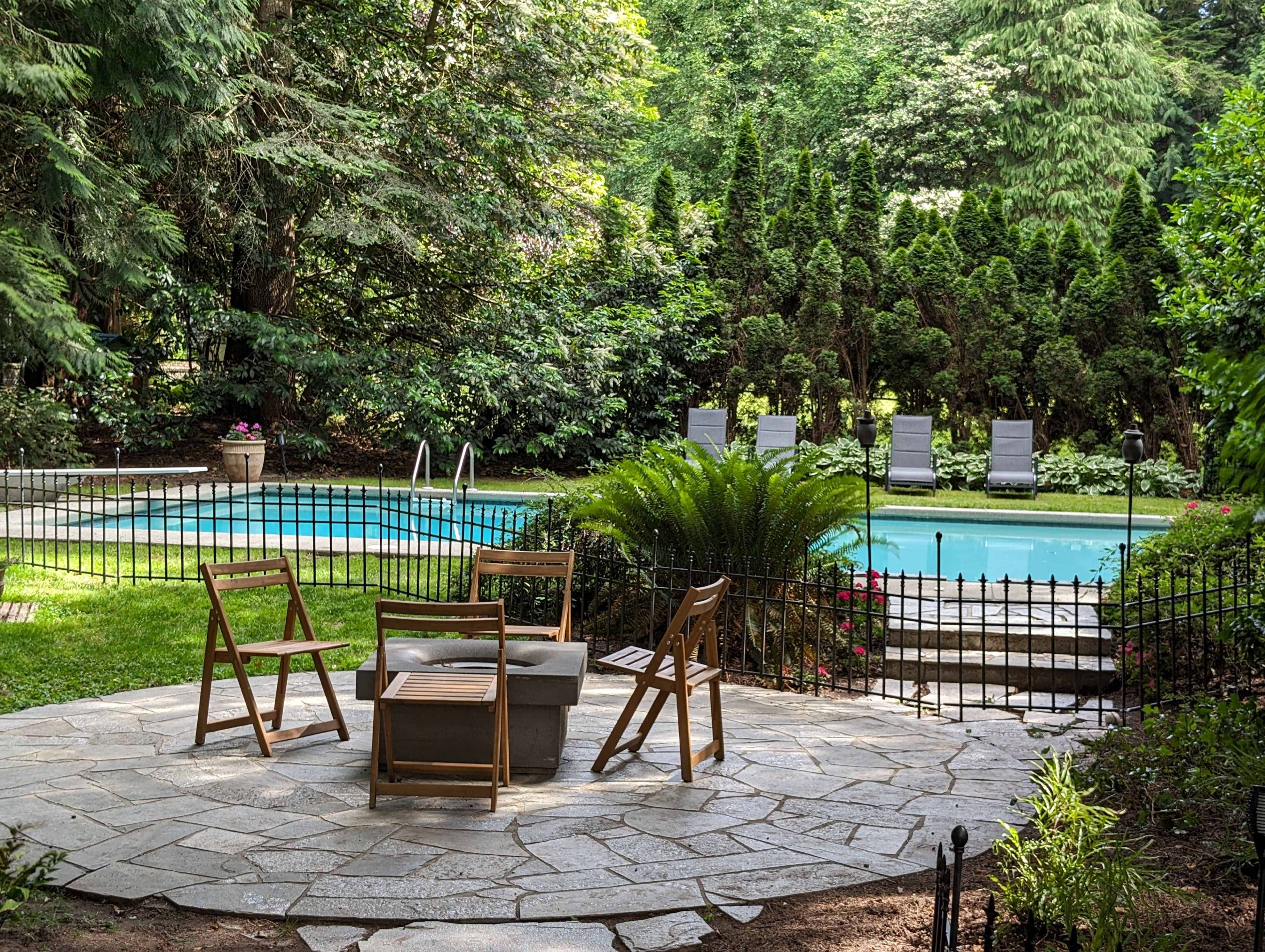 A stone patio with wooden chairs and a small table overlooks a swimming pool surrounded by greenery and lounge chairs.
