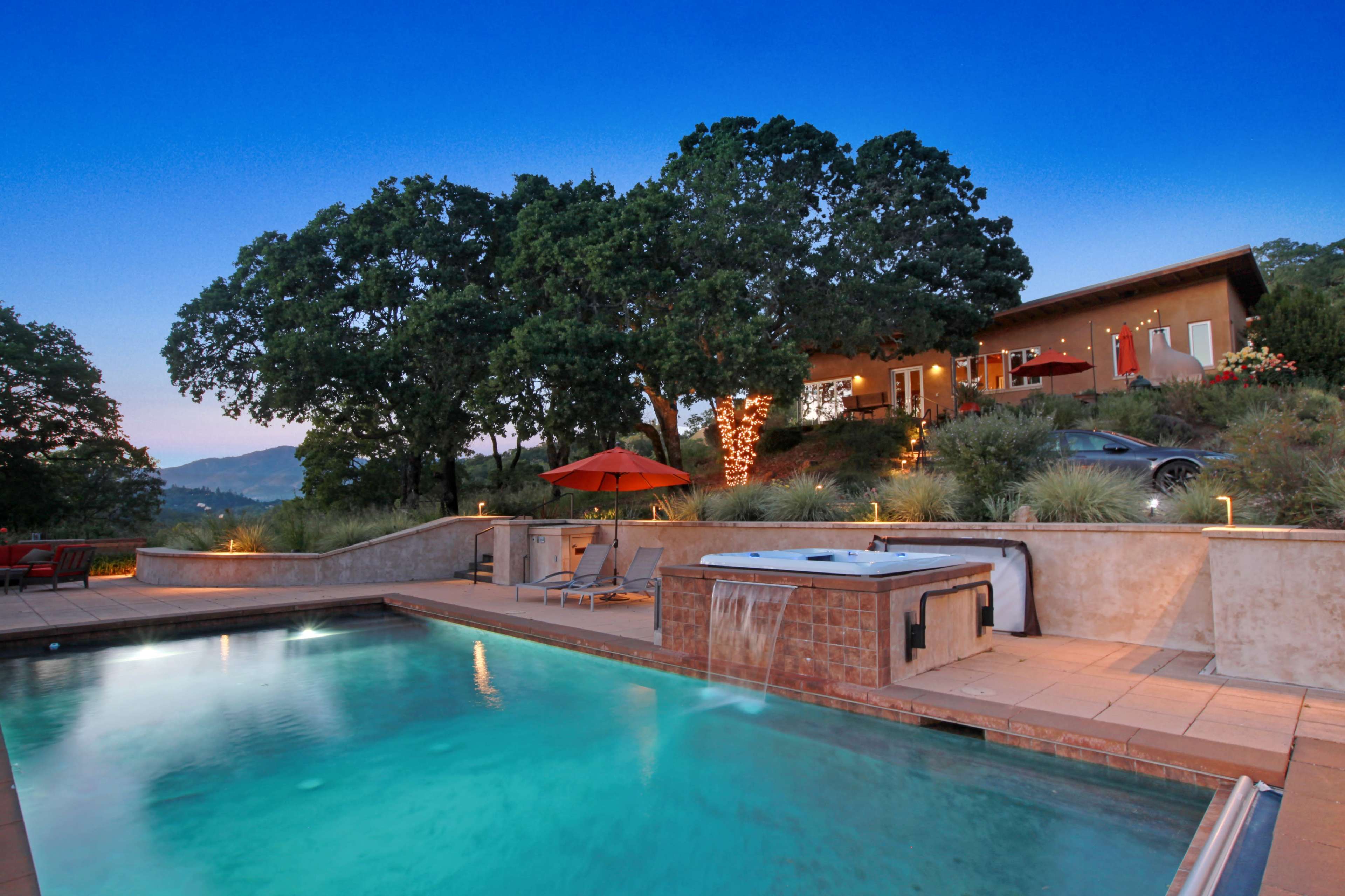 A modern home with large windows overlooks a swimming pool and hot tub, surrounded by trees and landscaping at dusk.
