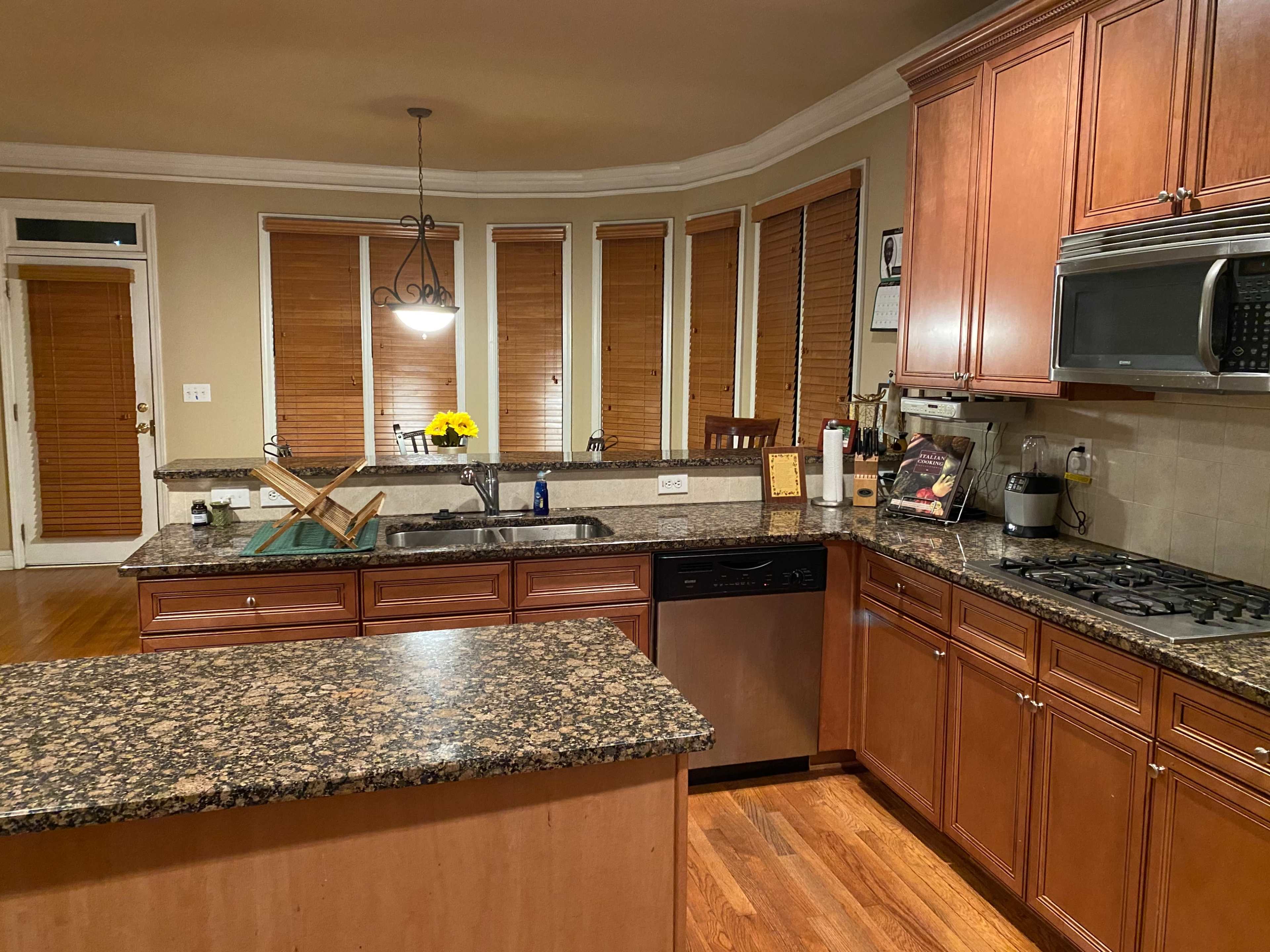 A modern kitchen with wooden cabinets, a granite countertop, and several windows covered by blinds.