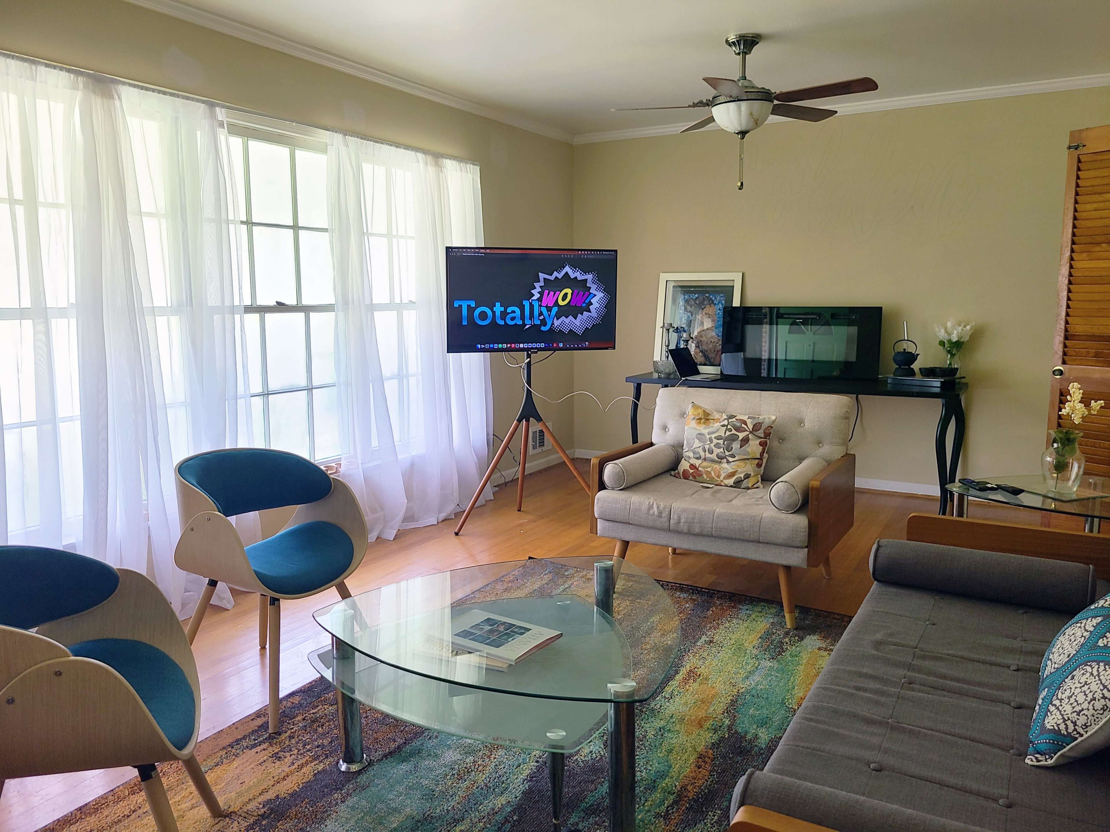 The image shows a well-lit living room featuring a large television on a stand, a glass coffee table, several chairs, and a sectional sofa.