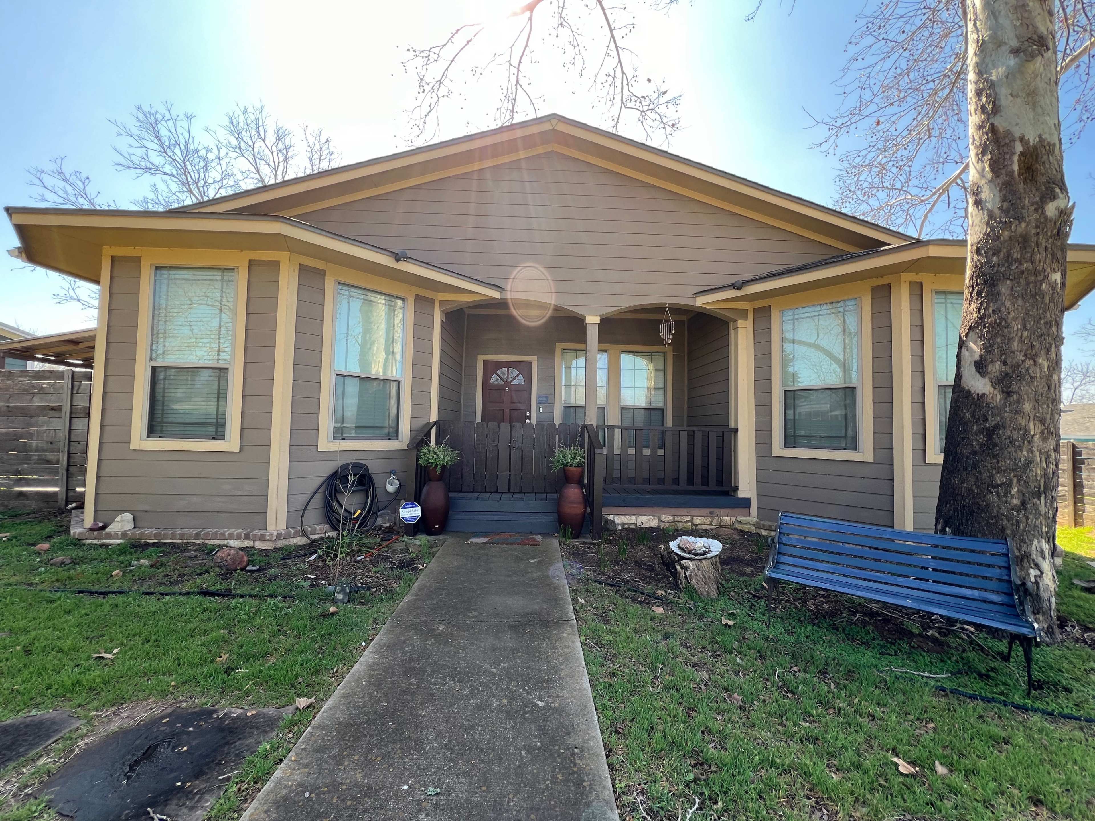 A single-story house with a front porch, large windows, and a pathway leading to the entrance is surrounded by a lawn and a bench.