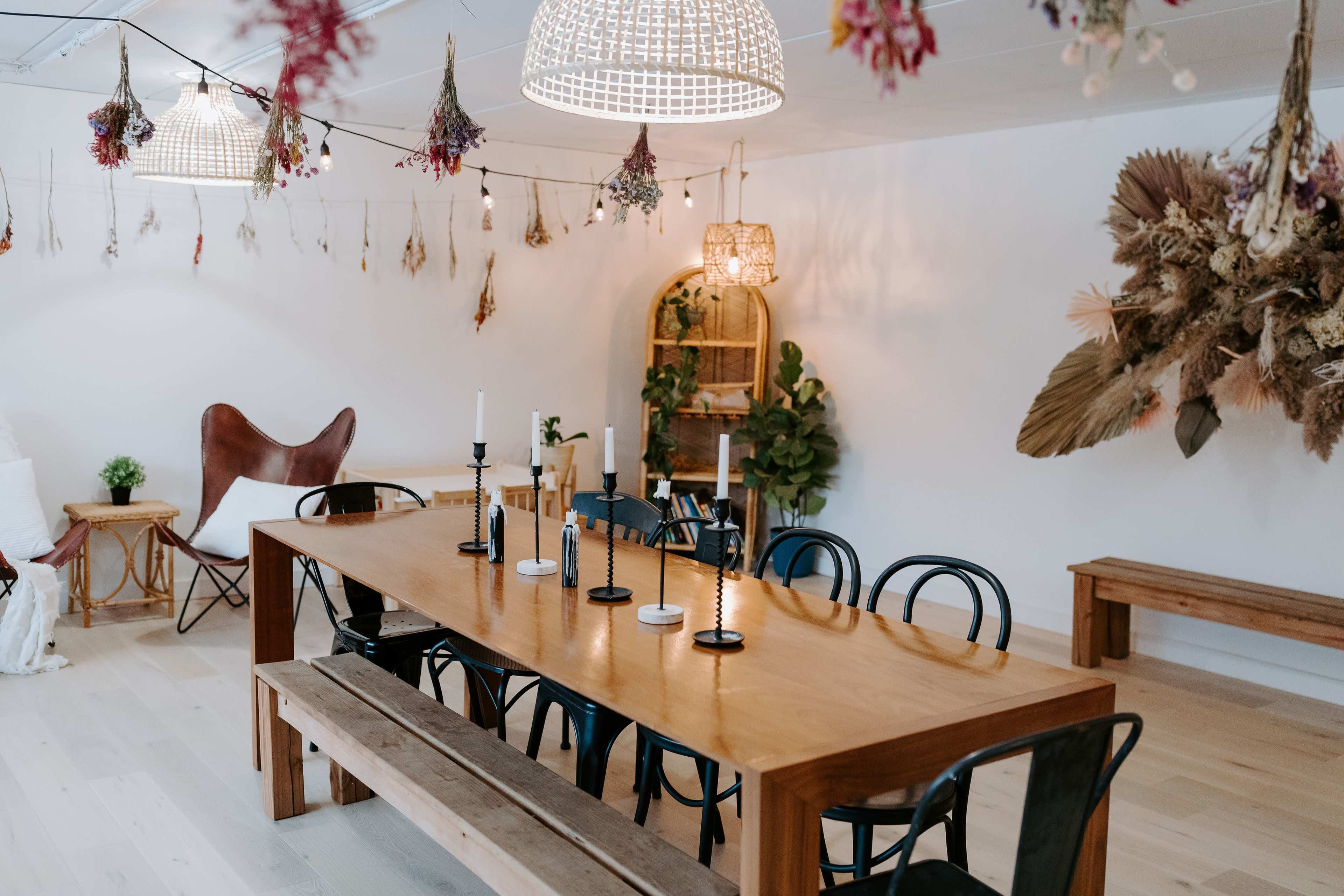 A spacious dining area features a large wooden table surrounded by black chairs and decorated with candles, while hanging plants and dried flowers adorn the walls and ceiling.