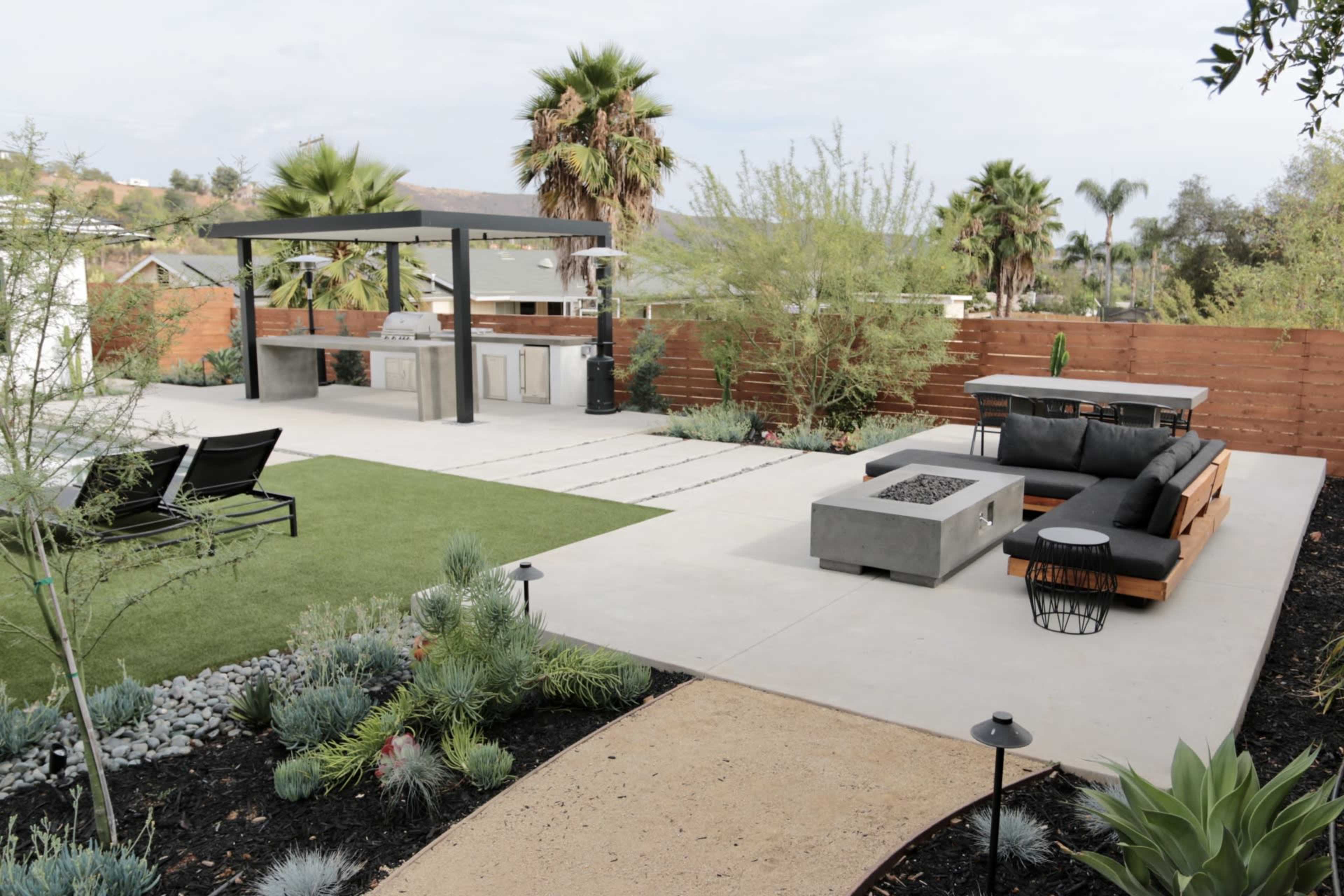 The image shows a modern backyard featuring a concrete patio with a fire pit, lounge seating, an outdoor kitchen, and landscaped greenery.