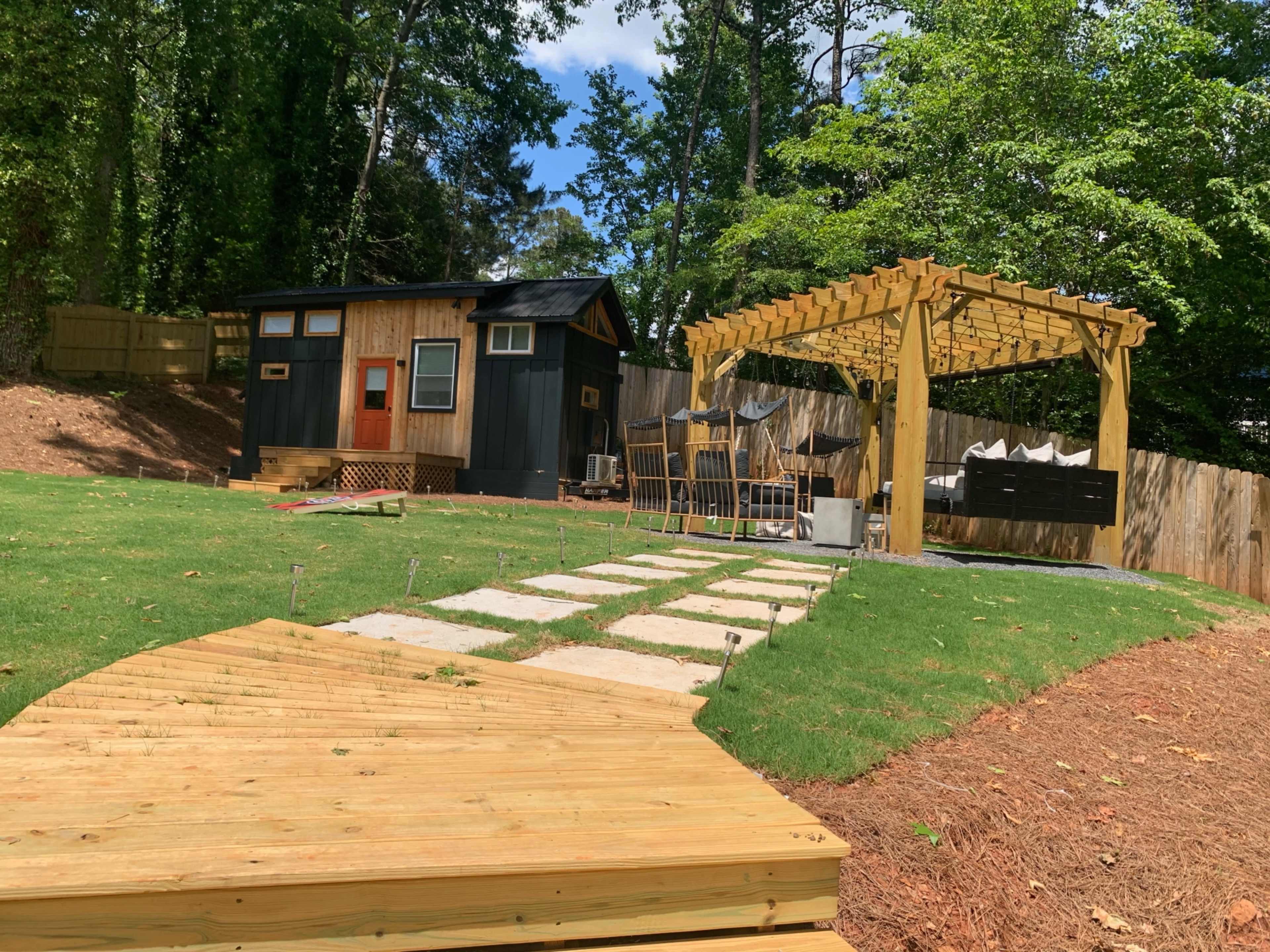 A small dark-colored cabin sits in a backyard with a wooden pergola, patio furniture, and a stone pathway leading through the grass.