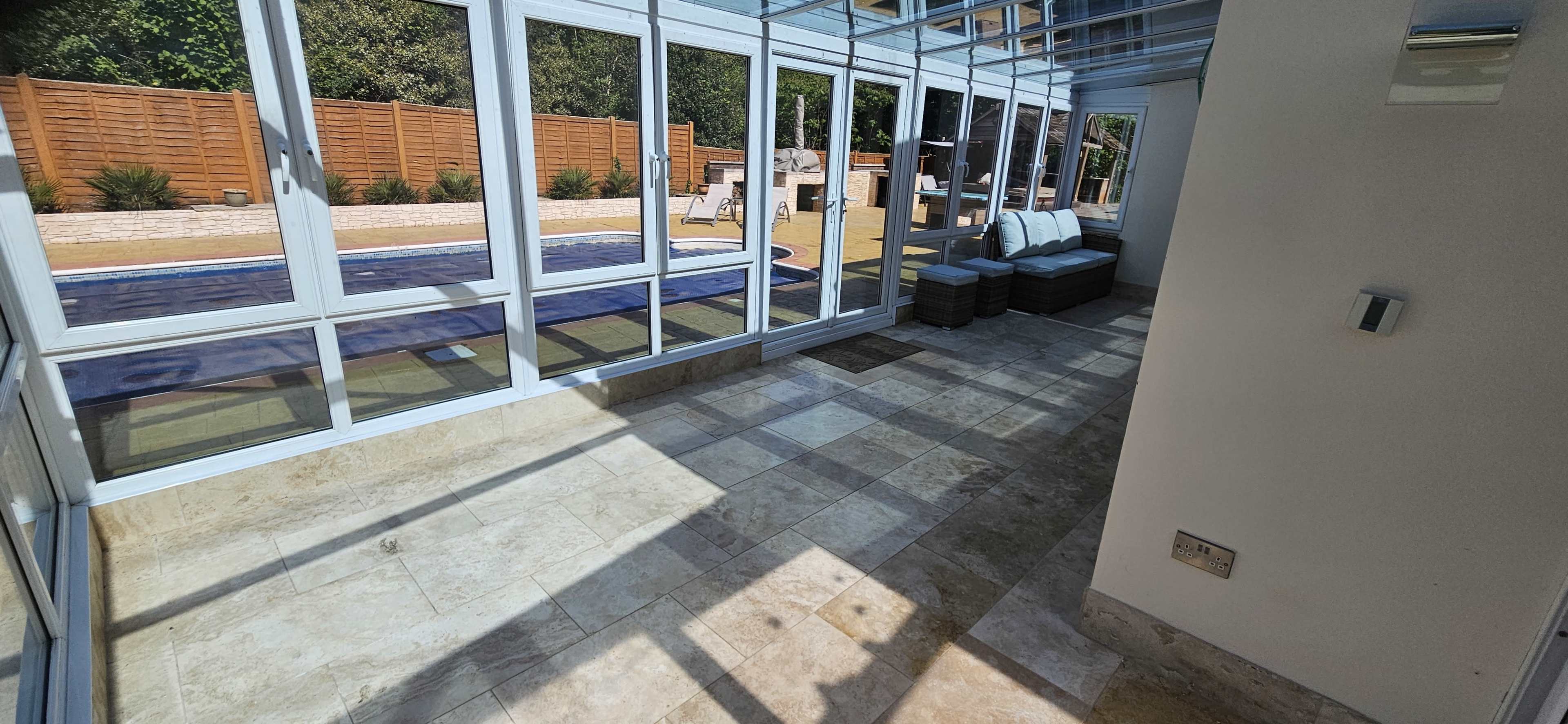 The image shows a sunlit room with large glass windows overlooking a patio and swimming pool, featuring tiled flooring and a seating area with cushions.