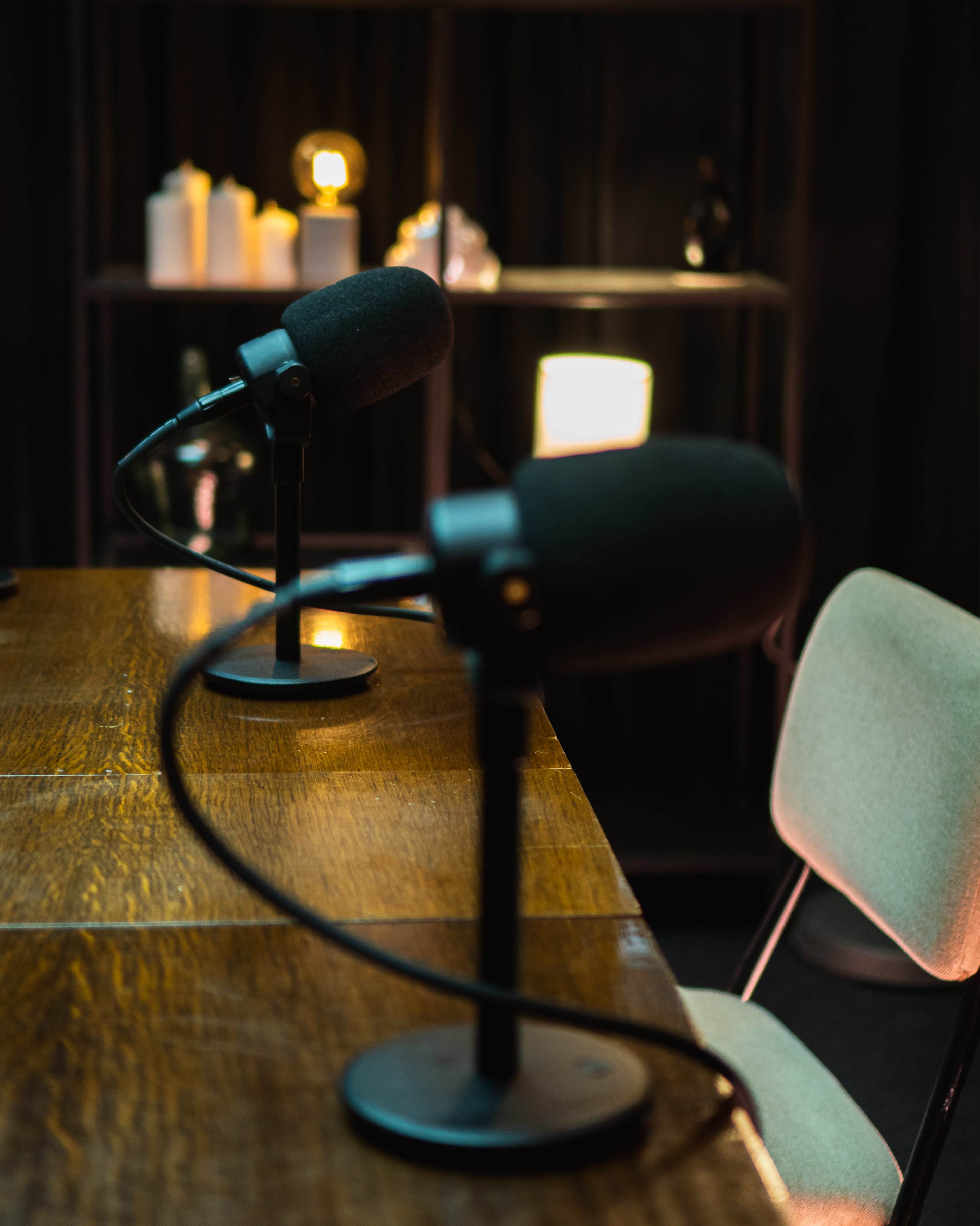The image shows two microphones positioned on a wooden table, with a blurred background of lights and shelves.
