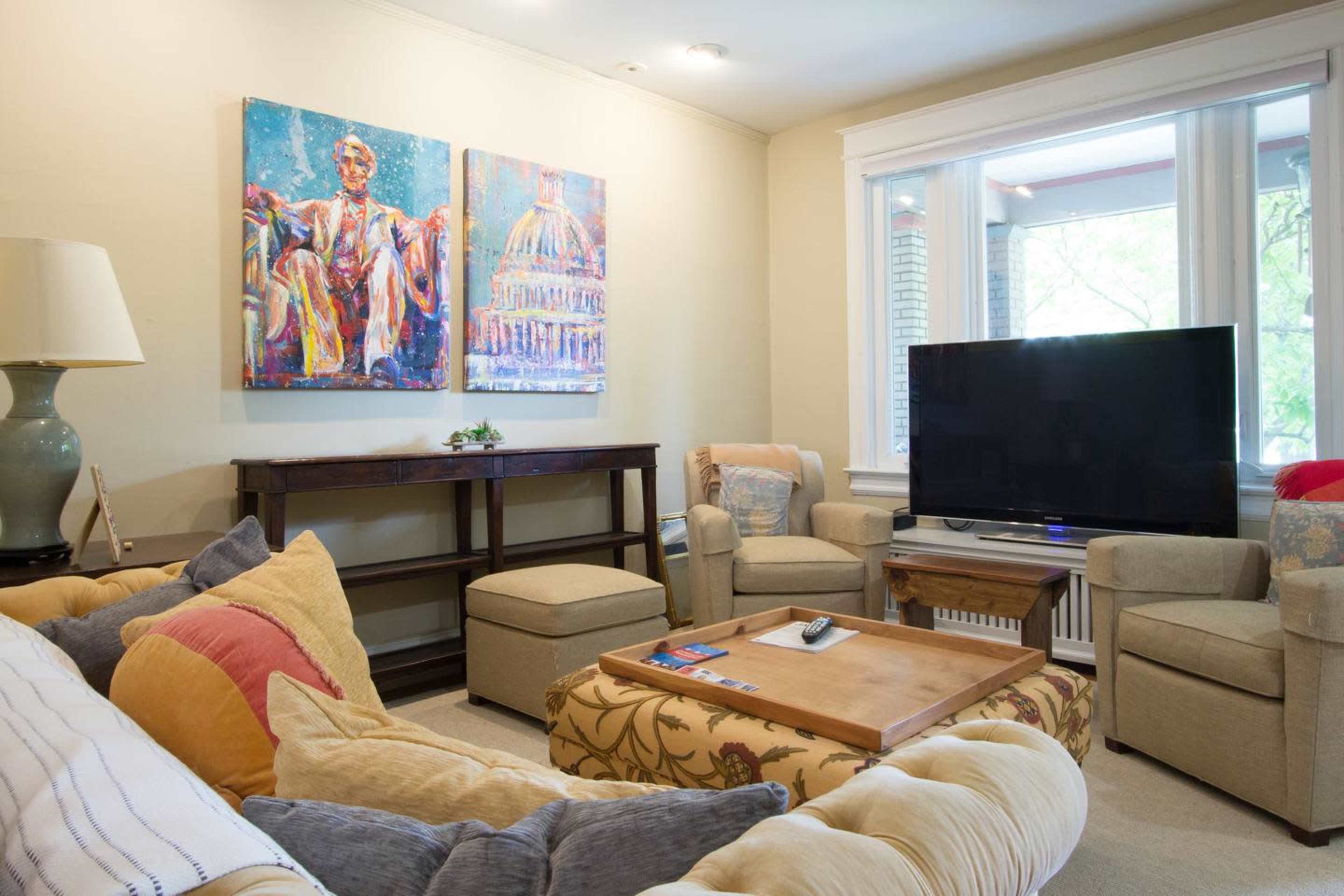 A cozy living room featuring two colorful paintings, a large television, a wooden coffee table, and comfortable seating arrangements.