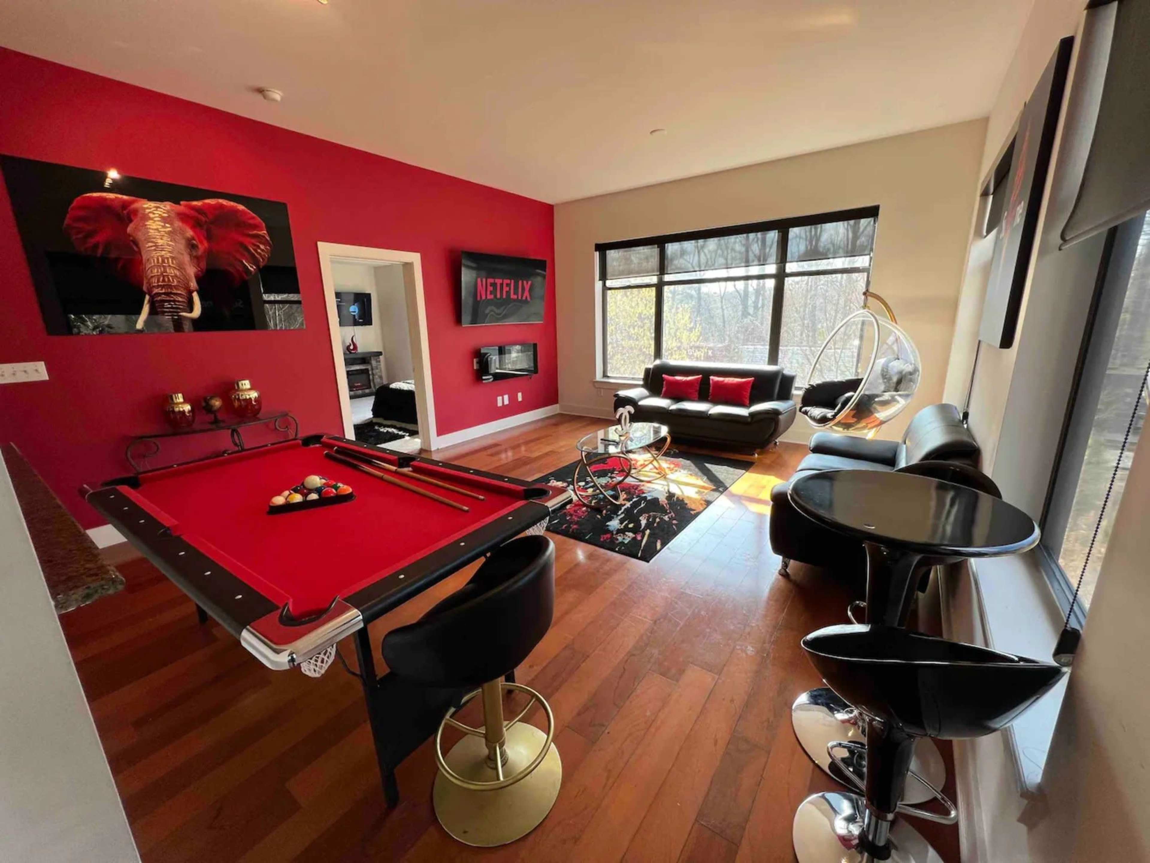 A modern living room with a pool table, a black leather sofa, and large windows overlooking a wooded area.