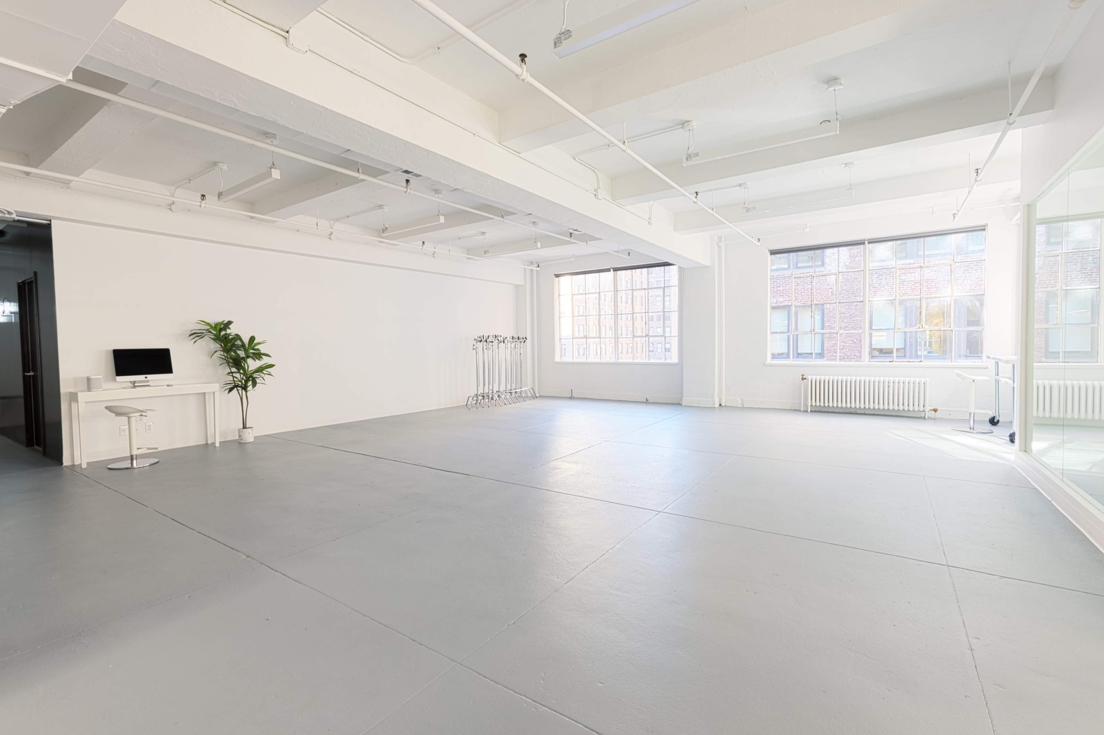 A spacious, well-lit studio with gray flooring, large windows, and minimal furniture including a desk and a plant.