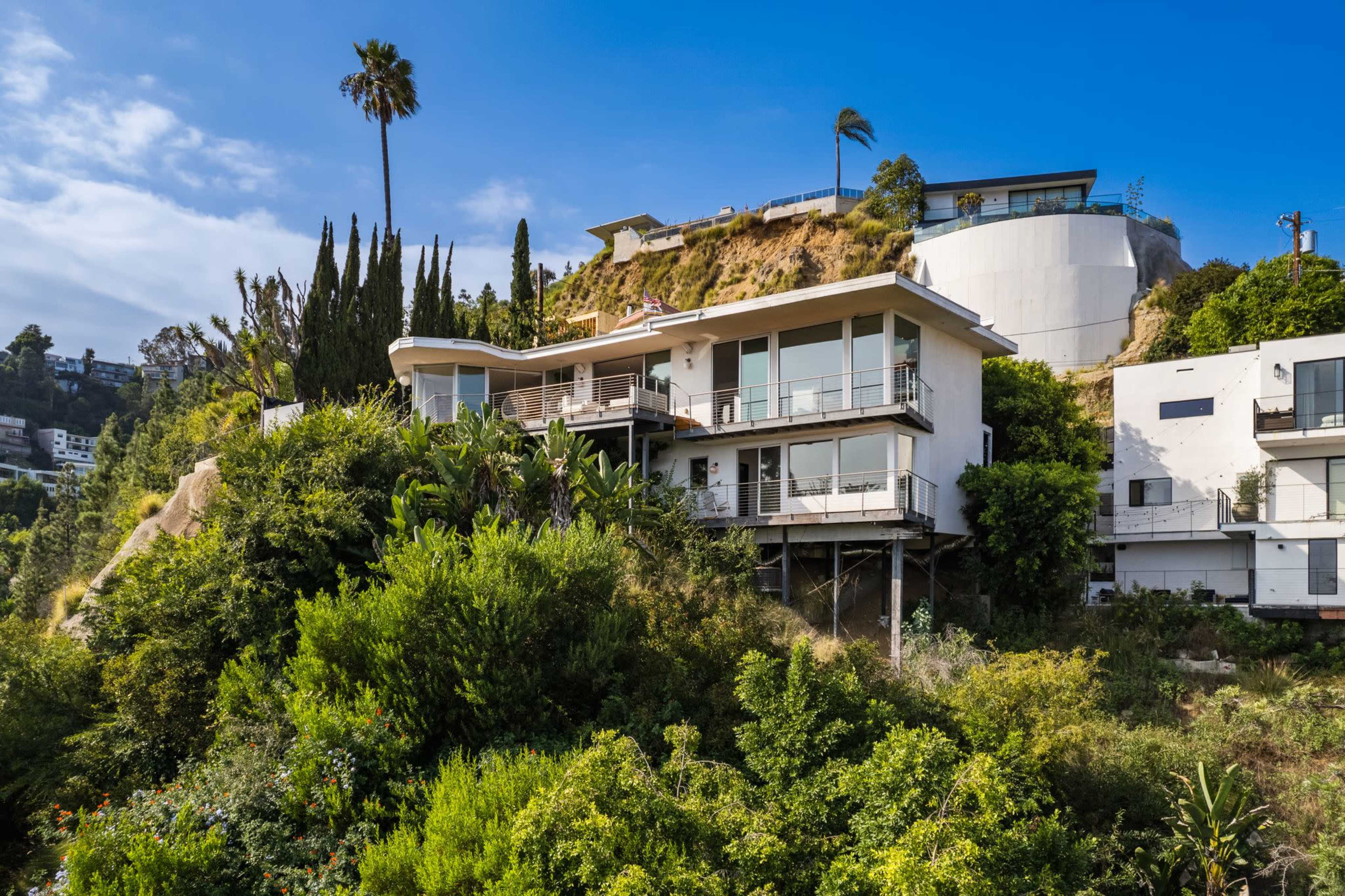 A modern house sits on a hillside, surrounded by lush greenery and tall trees, with a clear blue sky above.