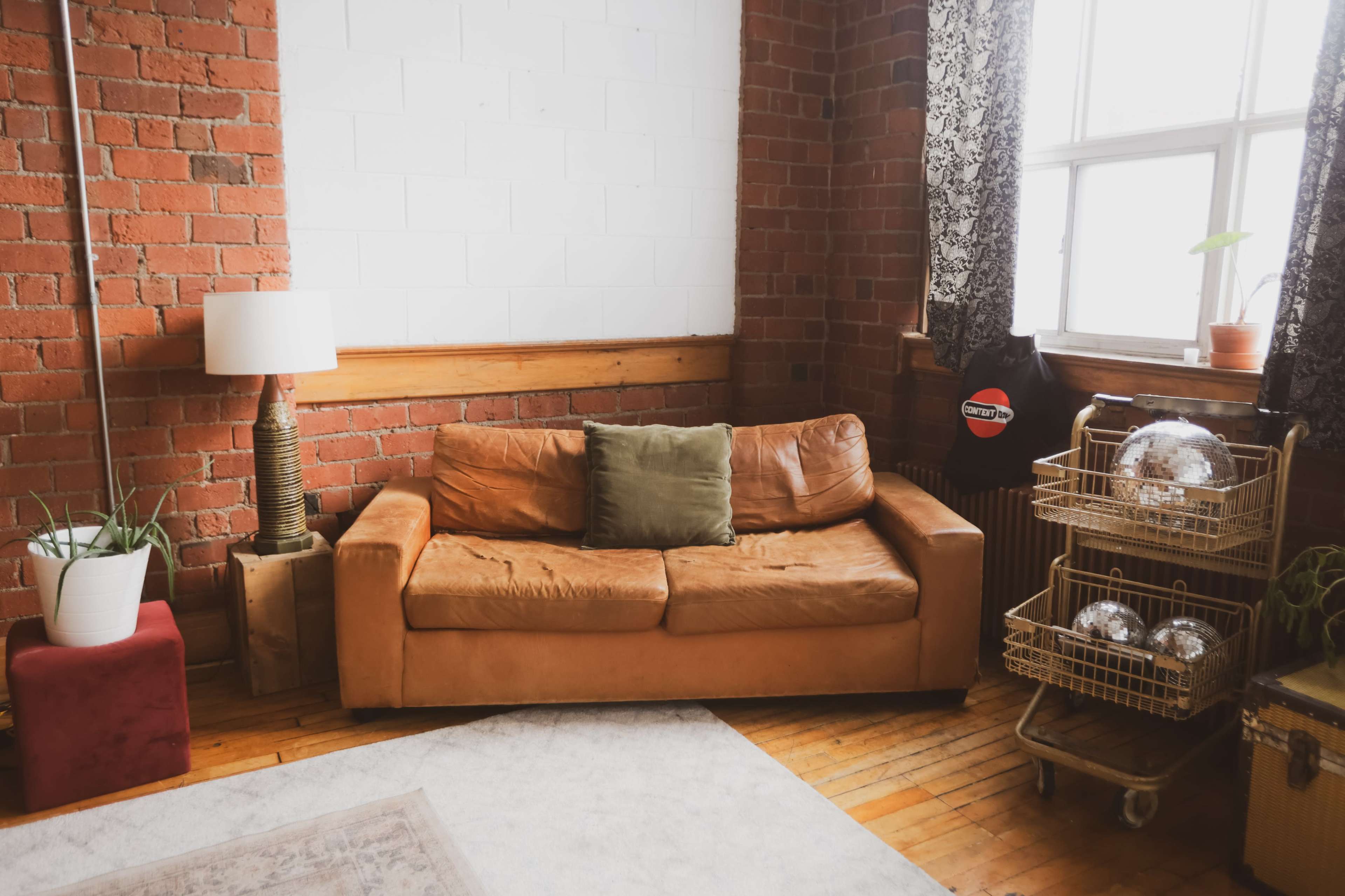 A brown leather sofa is positioned against a brick wall, accompanied by a lamp and various decorative items, including plants and a cart with metallic spheres.