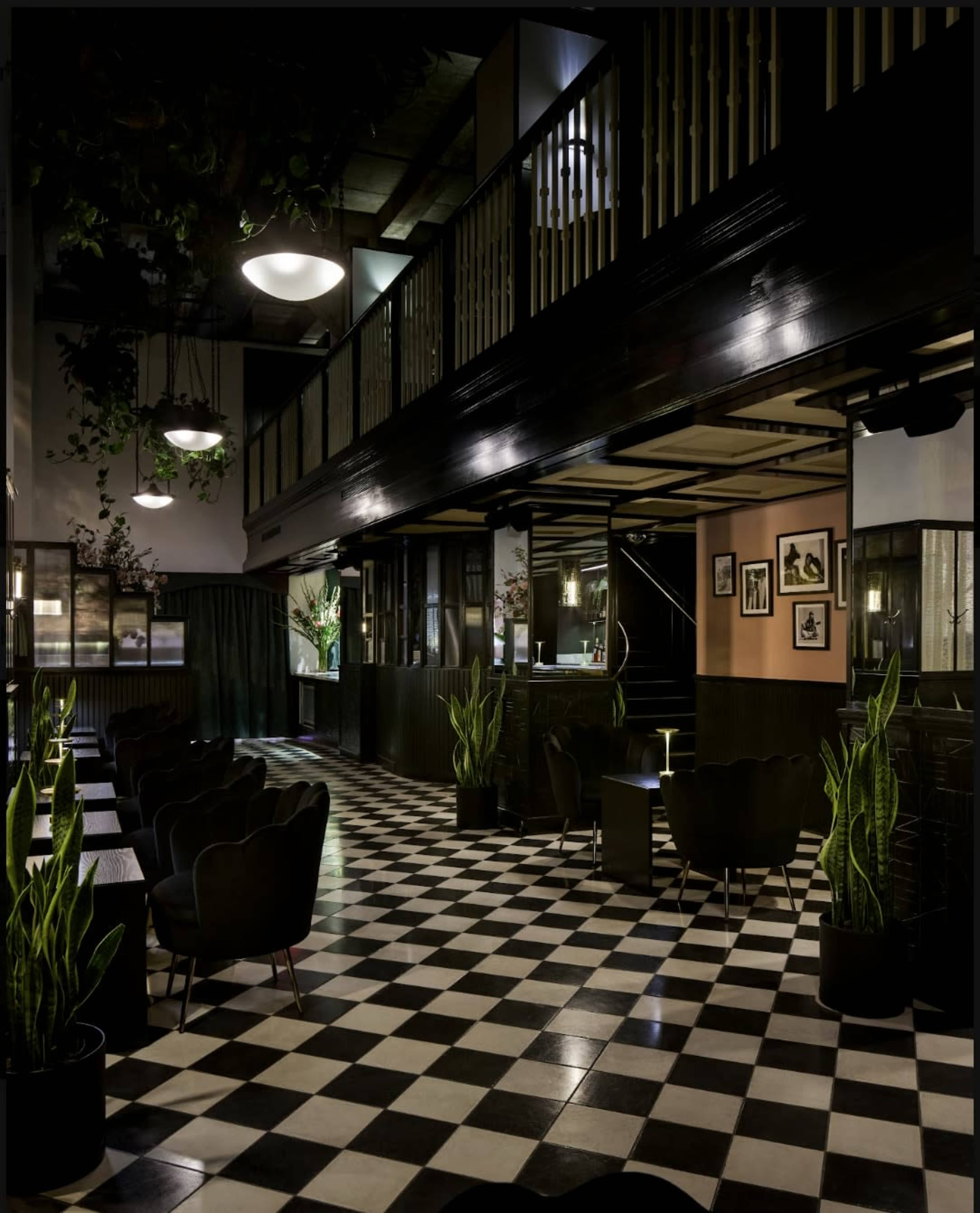 The image shows a stylish interior of a café or lounge with a black and white checkered floor, potted plants, and seating areas illuminated by pendant lights.