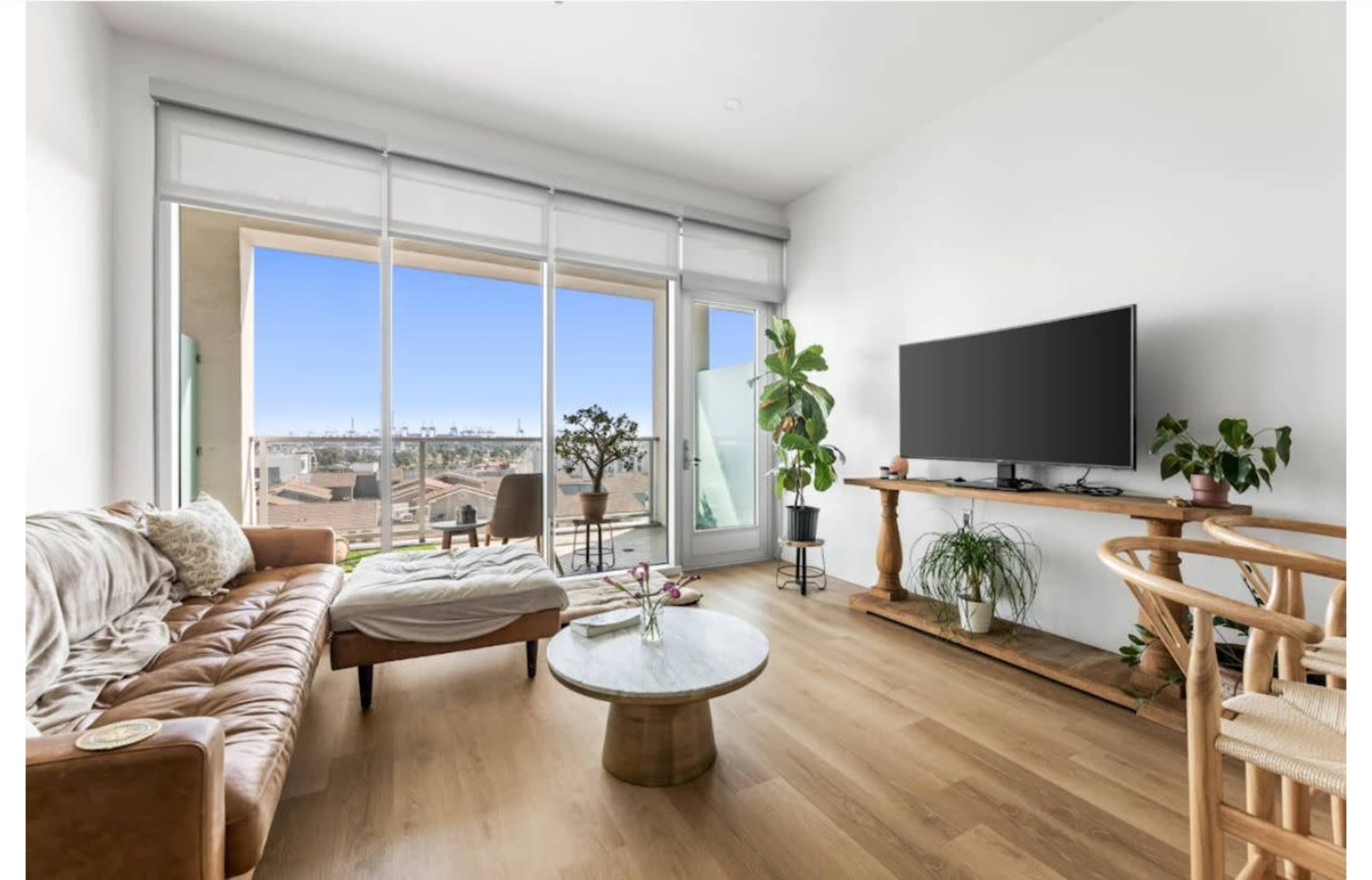 The image shows a modern living room with a brown leather sofa, a circular coffee table, a television on a wooden stand, and large windows providing a view of the outdoors.