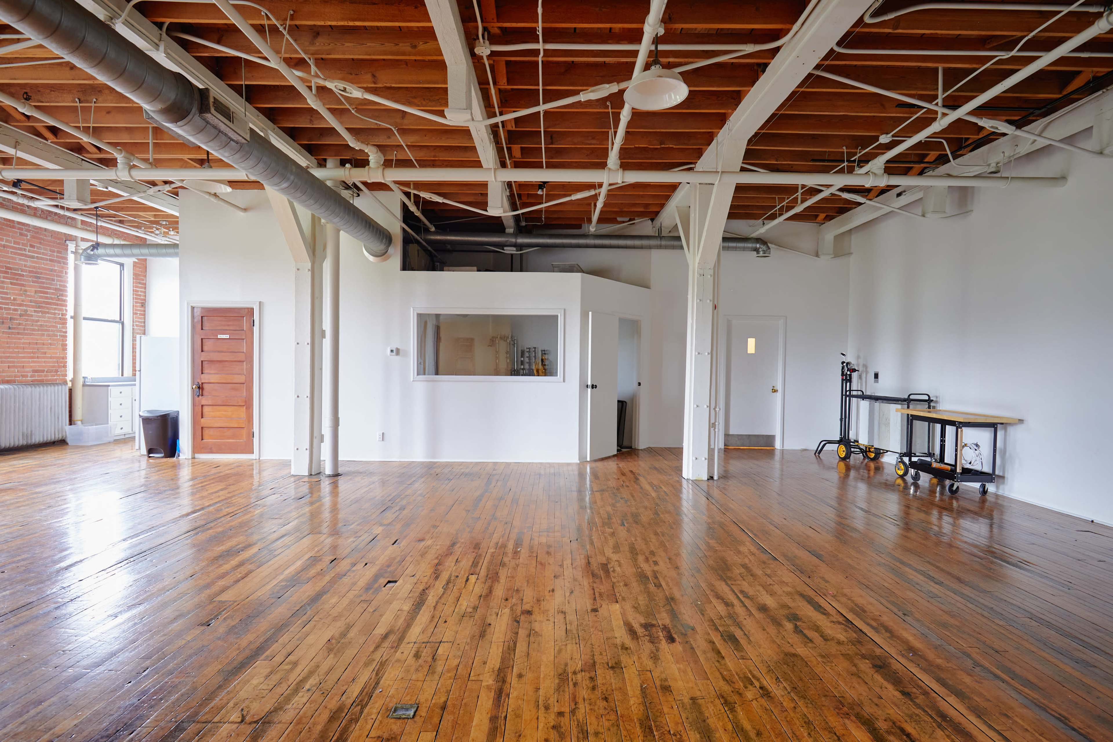 A spacious room with wooden floors, exposed beams, and a large window, featuring an area separated by a curved partition and a doorway leading to another room.